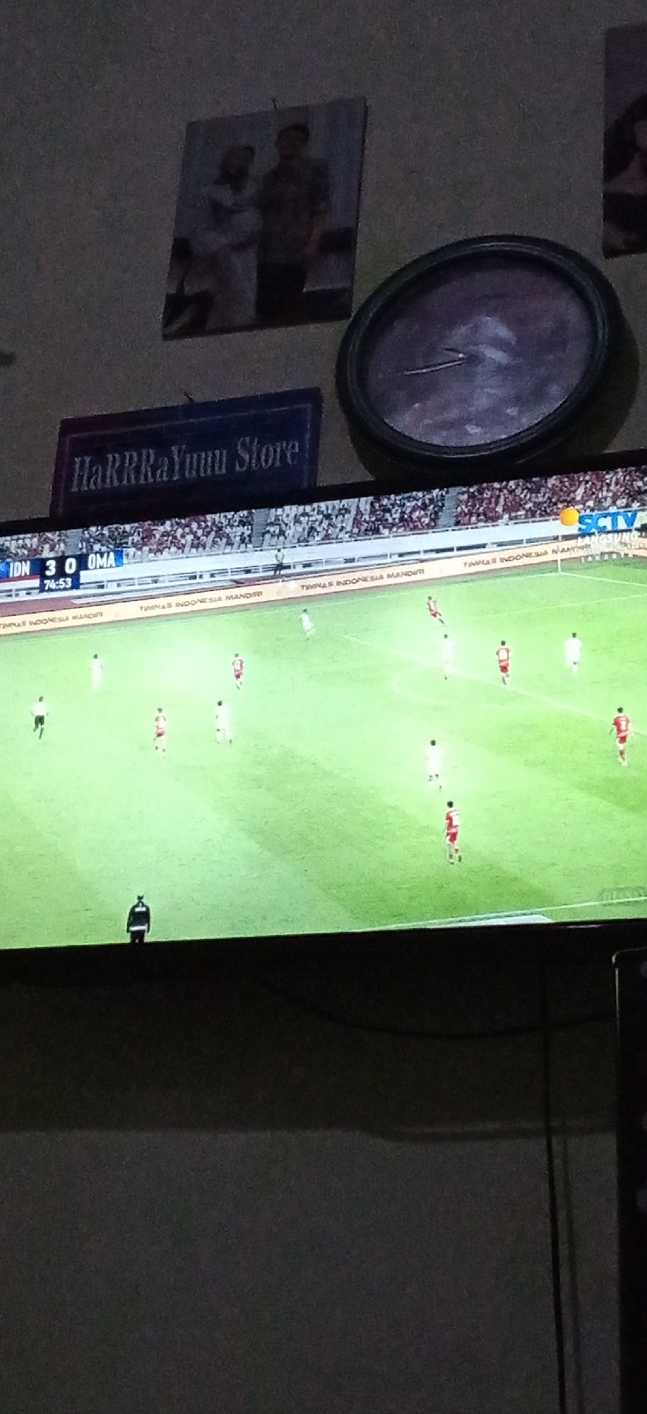 The image presents a dimly lit indoor scene in the city of Banjaran, Indonesia, focused on a television displaying a football (soccer) match. The score overlay indicates "IDN 3 0 OMA" (Indonesia leading Oman) at "74:53" (74 minutes, 53 seconds). The broadcast logo "SCTV" is visible in the upper right corner of the screen, and a sponsor banner reading "TIMNAS INDONESIA MANDIRI" is visible along the perimeter of the virtual stadium. Multiple players in red and white jerseys are visible on a green pitch, with one figure in a black jacket standing near the bottom left edge of the television screen.
Above the television, mounted on a light-colored wall, are several objects. From left to right: a framed picture depicting two individuals, a man and a woman in a headscarf; a dark blue rectangular sign with white text that reads "HaRRRaYuuu Store"; and a circular wall clock with a dark frame and a dark face, showing visible hands. A partial view of another framed picture is visible on the far right. The area below the television is dark, with visible cables. The primary light source appears to be the television screen itself.