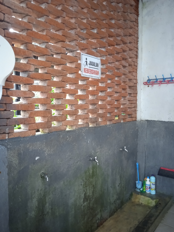 The image shows a public wash area in the city of Plaosan, Indonesia, at the coordinates -7.669, 111.217. The area is a small, open-air space with a concrete floor and a brick wall. Two faucets protrude from the wall, with the left one turned off and the right one turned on. A blue brush leans against the wall next to the right faucet, and a blue and white bucket sits below it. Two bottles of cleaning liquid sit on a small concrete shelf below the faucets. The wall above the faucets is made of red bricks laid in a staggered pattern. A sign with the text “Jagalah kebersihan” is affixed to the wall, which translates to "Maintain cleanliness" in English. The wall to the right of the wash area is made of white concrete. A blue and red clothesline hangs from this wall. The entire scene is dirty, with green algae growing on the walls and concrete floor. The image is taken from a low angle and is in focus. There is no one in the photo.