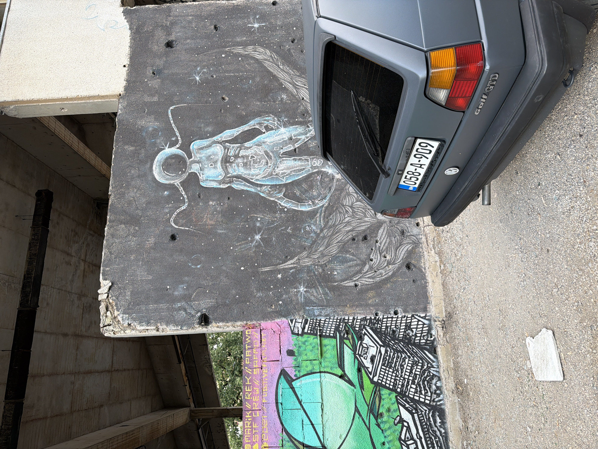 In Mostar, Bosnia and Herzegovina, a gray Volkswagen Golf GTD is parked on a gravel lot in front of a graffiti-covered concrete wall. The wall features a stylized blue humanoid figure with radiating lines, set against a dark background with white specks resembling stars. To the left of this is a section of a brick wall with vibrant graffiti depicting a large green sphere and urban buildings. The background suggests an abandoned or under-construction building with visible concrete beams and unfinished ceilings. The car's license plate, "058-A-909", is clearly visible. The overall scene depicts urban decay and artistic expression.