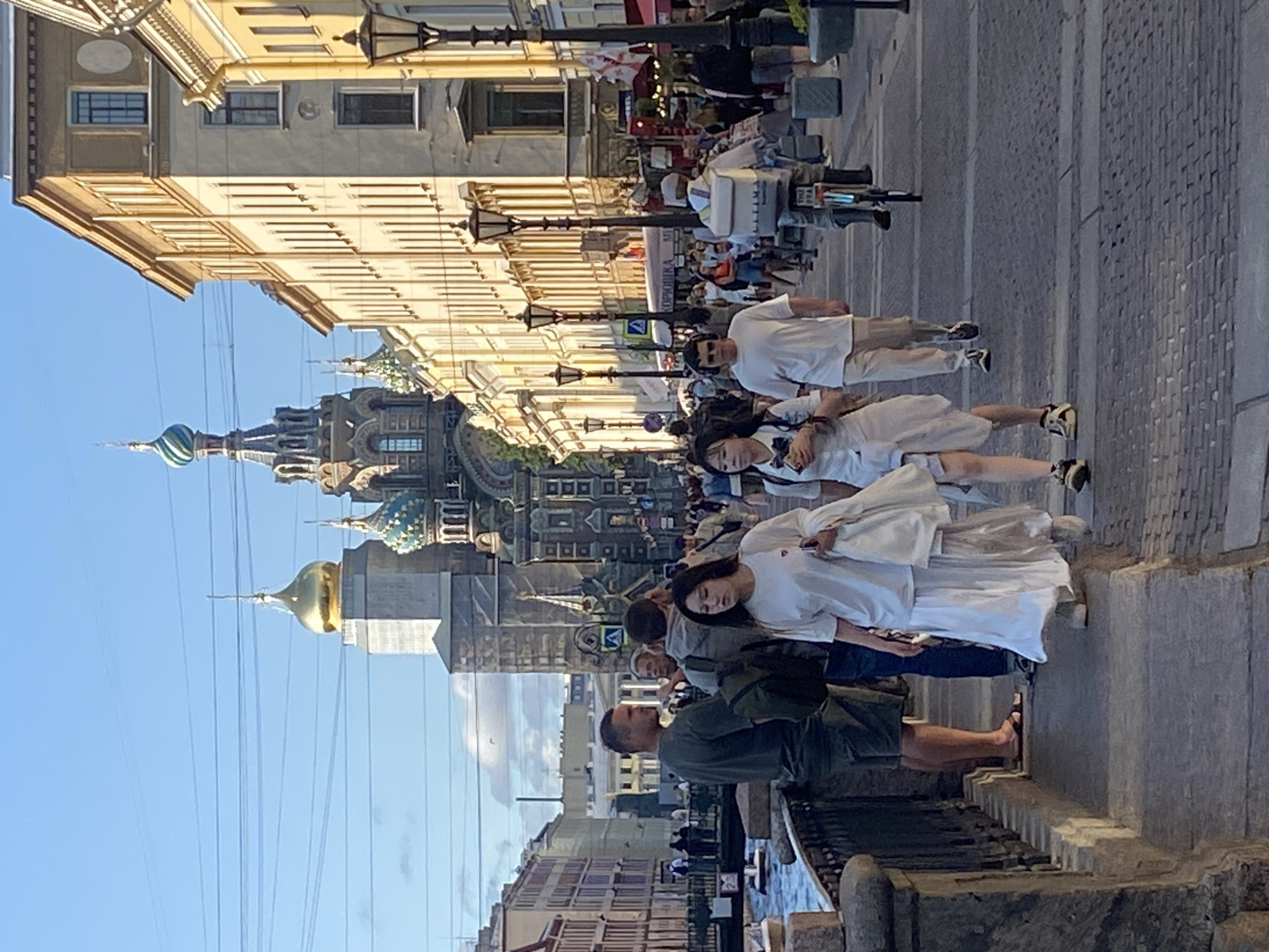 The image depicts a street scene in Saint Petersburg, Russia, likely on Nevsky Prospekt, given the presence of the Church of the Savior on Spilled Blood in the background, a prominent landmark. The sun is casting long shadows, indicating it's either morning or late afternoon. Several people are walking along a wide, paved walkway bordering a canal.
In the foreground, two women are walking towards the viewer. The woman on the left is wearing a white t-shirt and a long, flowing white skirt. The woman on the right is wearing a white t-shirt and white shorts. They appear to be together, with the woman on the left carrying a white bag over her shoulder.
Behind them, a man in a dark green t-shirt and shorts is standing, looking to his left. Next to him, a group of people are gathered, engaged in conversation. Further down the walkway, a man is cycling with a large white delivery box attached to the back of his bicycle. Other pedestrians are visible in the middle ground and background, some casually strolling, others appearing to be in conversation.
The architecture lining the street features classical European-style buildings with multiple stories, adorned with windows and decorative elements. Lampposts are spaced along the walkway. The canal on the left is bordered by a stone embankment and a metal railing. The sky is clear blue with a few wispy clouds. The overall atmosphere is one of a busy, picturesque urban environment.