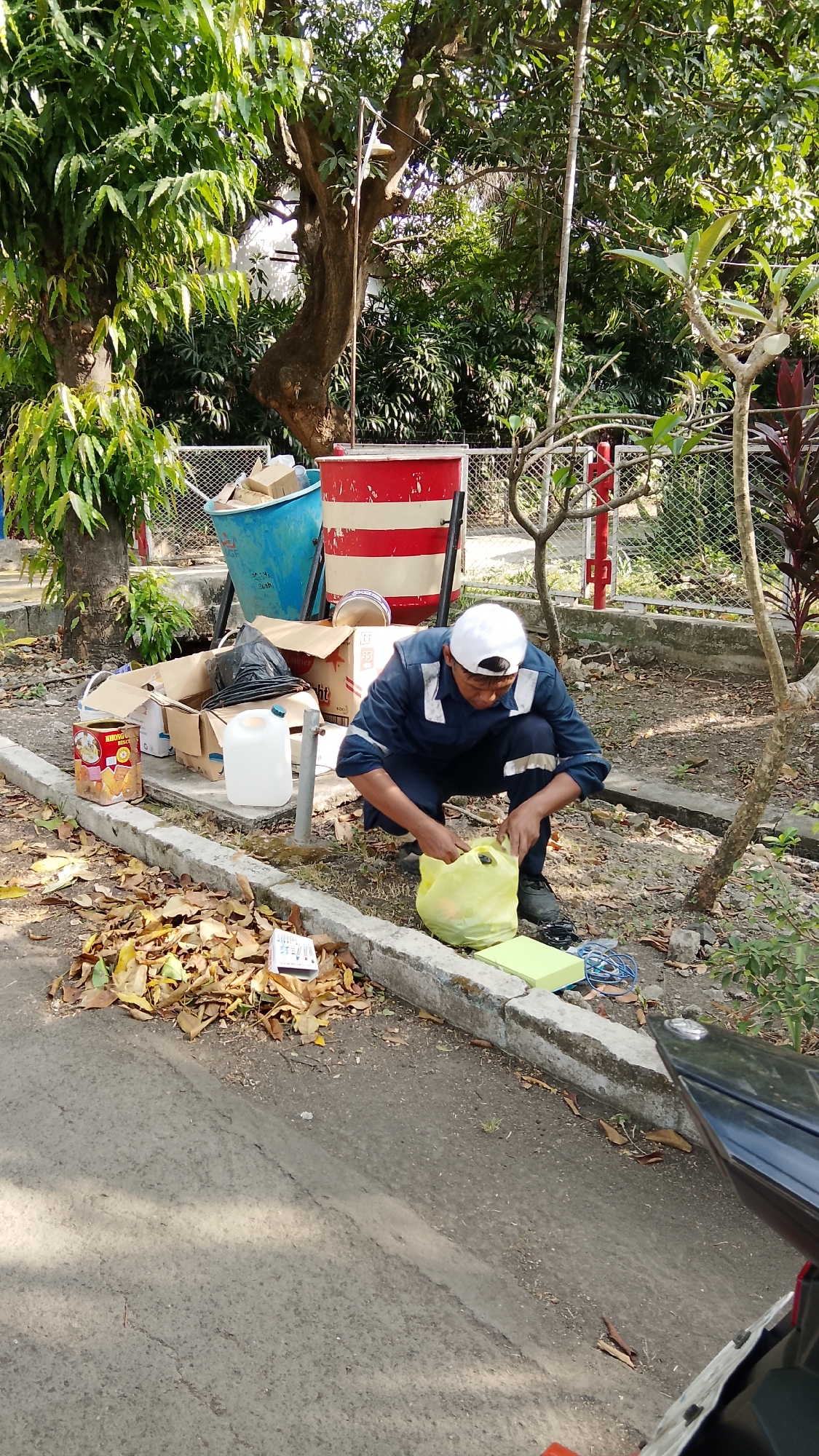 In Tlogobendung, Indonesia, a man wearing a blue uniform and a white cap is crouching beside a pile of discarded items. The items include cardboard boxes, a blue bin, a red and white striped bin, a white jug, and a yellow plastic bag. The man is interacting with the yellow plastic bag. The scene appears to be an outdoor area with trees and a fence in the background. There is a pile of fallen leaves on the ground near a curb. A portion of a black motorcycle is visible in the lower right corner of the image.