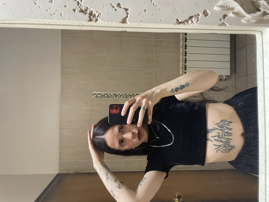 A mirror selfie depicts a female subject inside a room, likely a bathroom, in Ushuaia, Argentina. The subject has long dark hair, dark red lipstick, and a small ring piercing in her left nostril. She wears a black, short-sleeved velvet crop top and dark-colored pants, revealing her midriff with a navel piercing. Visible tattoos include an intricate design on her left forearm/upper arm, a stack of four stylized symbols (one blue) on her right forearm, and a large black abstract or stylized flame design below her navel. She wears two silver chain necklaces, one with a small "I" pendant, and a ring on her right ring finger. Her right hand holds a black smartphone with a red camera module, positioned for the photograph, while her left hand rests on the top of her head with fingers slightly splayed. The background features light beige rectangular wall tiles, a vertical accent strip of smaller mosaic tiles, a dark wooden door with a gold-colored doorknob, and a white panel radiator. The ornate, textured mirror frame with relief patterns is visible on the right side.