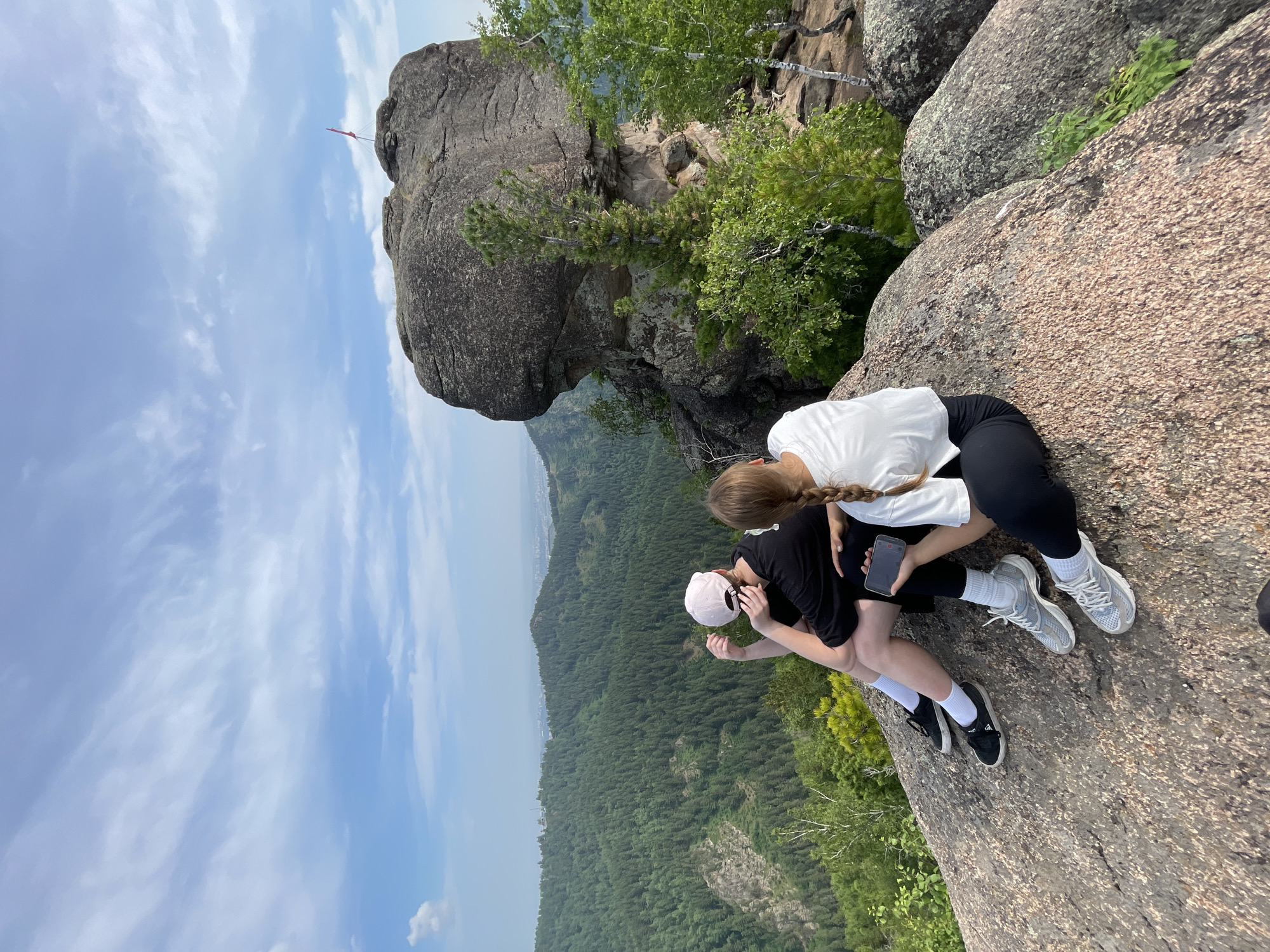 The image shows two people sitting on a rocky cliff edge, overlooking a vast expanse of evergreen forest. The sky is a pale blue with wispy white clouds, suggesting daytime, likely late morning or early afternoon. The weather appears clear and pleasant for outdoor activities.
The subjects are two individuals, their backs to the camera, both dressed casually for an outdoor excursion. One person wears a white t-shirt and black leggings, while the other is in a black t-shirt, black leggings, and a light-colored baseball cap. They are positioned close together, with one holding a smartphone. Their posture suggests they are resting and taking in the view.
The setting is a dramatic, rocky outcrop with a sheer drop to the forested valley below. Large, weathered rock formations dominate the immediate foreground, with green trees and foliage clinging to the slopes and the distant landscape. The vastness of the forest and the clear sky create a sense of natural beauty and openness. A small red flag is visible on a distant rock formation, a subtle indicator of the location.
The overall mood of the image is one of peaceful contemplation and enjoyment of nature. The focus is on the scenic vista and the shared experience of being in a beautiful natural environment. There is no visible text in the image. The context provided places the scene in Krasnoyarsk, Russia, a region known for its natural landscapes.
