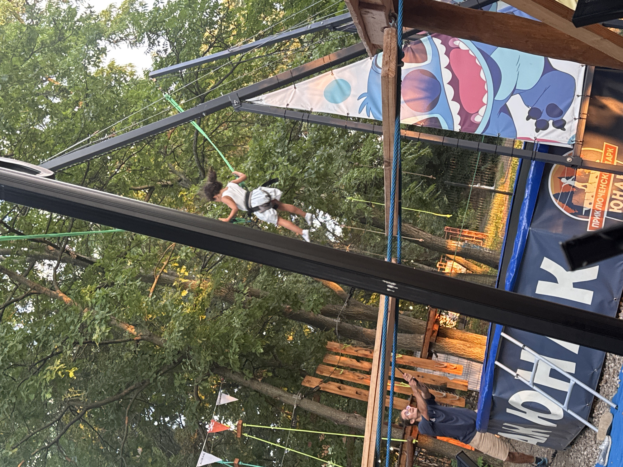 A young girl with dark curly hair is suspended in mid-air on a zipline. She is wearing a white dress and is harnessed to a green rope that runs along a metal track. She is positioned horizontally, with her arms outstretched and her legs slightly bent, as if in motion.
The setting appears to be an outdoor adventure park, with trees and greenery visible in the background. A large banner featuring a cartoon character, possibly Stitch from Disney's Lilo & Stitch, is hanging from a wooden structure. Another banner with Cyrillic text, which appears to be in Russian or Bulgarian, is also visible. The ground is covered with gravel.
A man with glasses is standing on the ground to the right of the frame, holding onto a rope and looking up. He is wearing a dark t-shirt and light-colored shorts.
The time of day appears to be late afternoon or early evening, as the sunlight casts long shadows and illuminates the trees. The weather seems pleasant, with clear skies. The overall mood of the image is one of excitement and adventure, with the girl appearing to be enjoying her ride on the zipline.