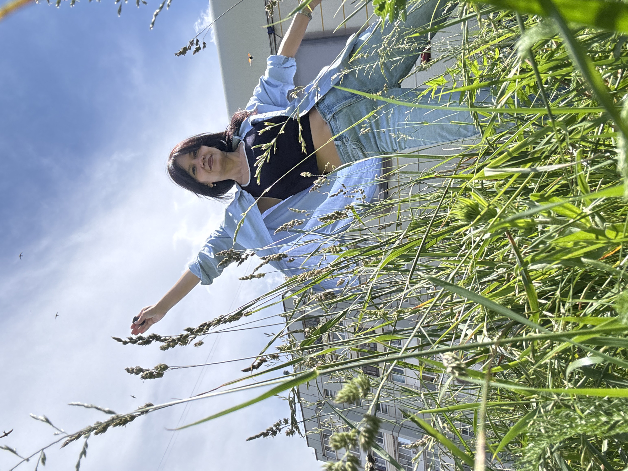 A woman with dark hair and fair skin, wearing a blue unbuttoned shirt over a black crop top and light blue jeans, stands in tall green grass. She is positioned in the foreground, facing forward, with her left arm extended upward and her right arm bent at the elbow, reaching towards the sky. Her left hand appears to be holding a small dark object. The background features a tall, light-colored building with multiple windows, indicative of a residential structure in an urban environment. The sky above is blue with scattered clouds, and several small birds are visible in flight. The perspective is low-angle, looking up at the woman and the building through the grass. The scene is set outdoors, under daylight conditions. The location is identified as Chaikovsky, Russia.