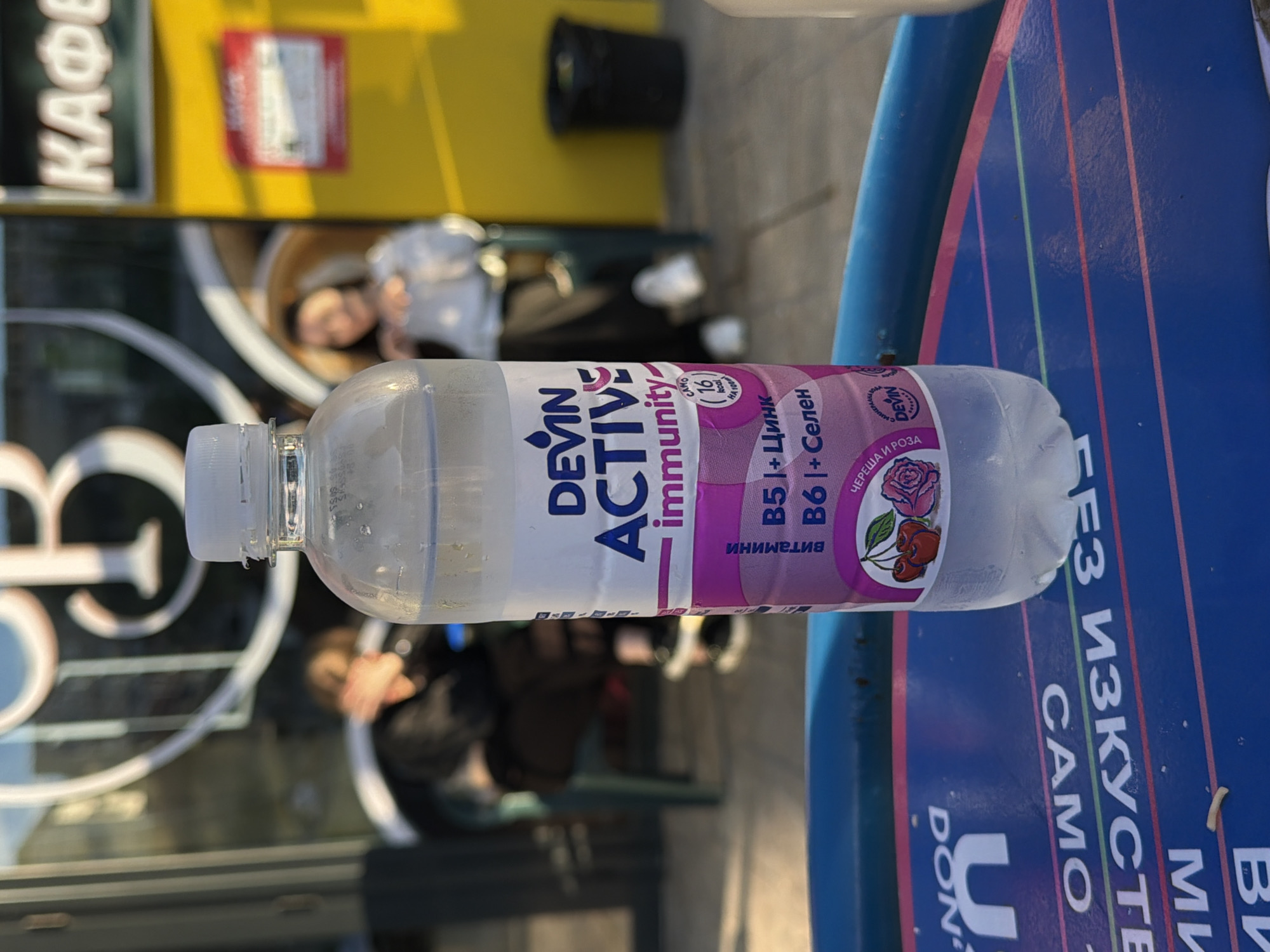 The image captures a clear plastic bottle of "DEVIN ACTIVE immunity" water, positioned on a blue table or surface in an outdoor setting in Shumen, Bulgaria. The bottle's label, featuring purple and white colors, indicates it contains "B5 + Цинк" (Zinc) and "B6 + Селен" (Selenium), with a flavor profile of "ЧЕРЕША И РОЗА" (Cherry and Rose), accompanied by icons of cherries and a rose. It also notes "16 kcal" and "витамини" (vitamins).
The background is brightly lit, suggesting daytime, possibly sunny. A blurred yellow structure, likely a building or kiosk, is visible behind the bottle, bearing a sign with the Cyrillic word "КАФЕ" (CAFE) in white letters. Another red rectangular sign is also visible on the yellow structure. Several blurred figures of people are in the background, one with dark hair appearing to be seated, indicating a public place like a cafe or street-side establishment. A black trash can is also discernible in the distant background.
The blue surface beneath the bottle has white and colored stripes and partial Cyrillic text, including "БЕЗ ИЗКУСТВЕ" (without artificial, likely part of a phrase like "без изкуствени оцветители" - without artificial colorants), "CAMO", "DEVIN", "DON", and "U". The overall scene suggests a casual moment, perhaps a break at an outdoor cafe or public area, with a focus on the branded immunity drink.