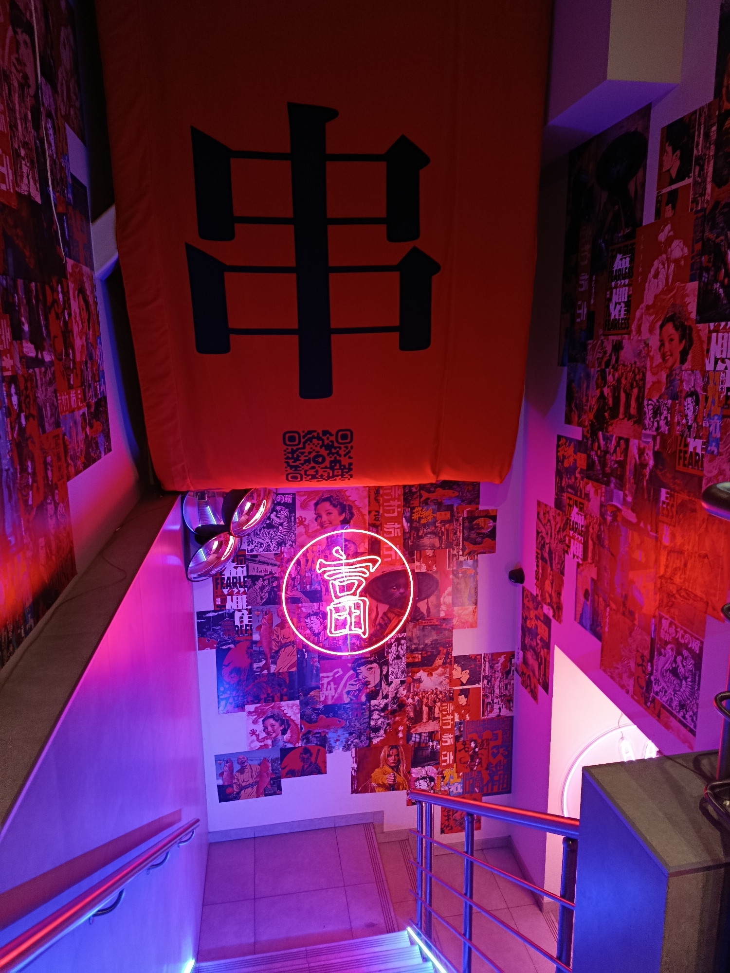The image depicts a narrow stairway with walls adorned with a collage of posters, predominantly featuring Asian cultural elements and imagery. A large orange banner with dark vertical markings hangs above the staircase, with a QR code visible on it. A neon sign in the shape of a circular motif with a stylized Chinese character, possibly representing "wealth" or "fortune," is prominently displayed on the wall. The stairway itself is illuminated by vibrant pink and blue neon lights, casting a colorful glow on the surroundings. The presence of a QR code and the general aesthetic suggest this could be a themed establishment, possibly a restaurant or bar, in Saratov, Russia. There are no individuals visible in the image.