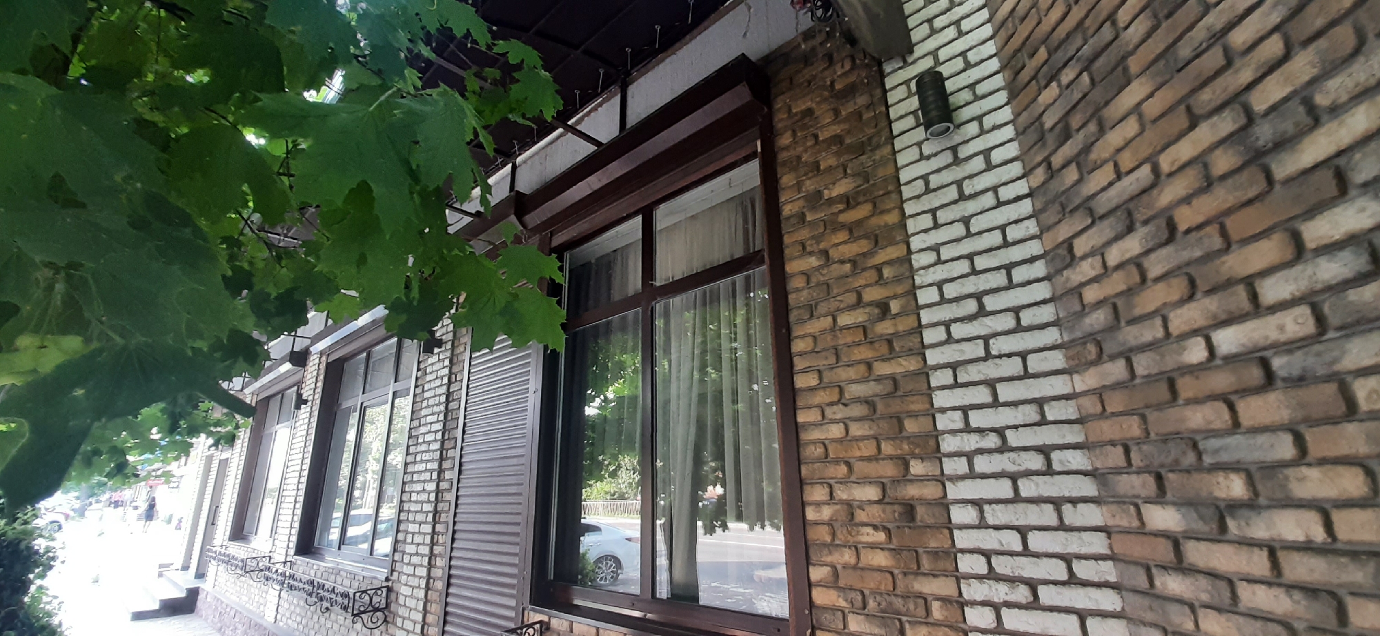 This media file shows the exterior of a building with large windows and a brick facade. The building appears to be a commercial establishment, possibly a cafe or restaurant, given the outdoor seating area and the presence of a street with vehicles and pedestrians in the background. The brickwork is a mix of light and dark brown bricks, with a section of white bricks forming a contrasting pattern. Green leaves from a tree in the foreground partially obscure the view of the building, suggesting it is a pleasant day with some foliage. The windows are framed in dark brown and have sheer curtains inside, offering glimpses of the interior. A black outdoor light fixture is mounted on the brick wall. The overall scene conveys a sense of a daytime setting in a town or city, with the architecture and street life typical of Yessentuki, Russia.