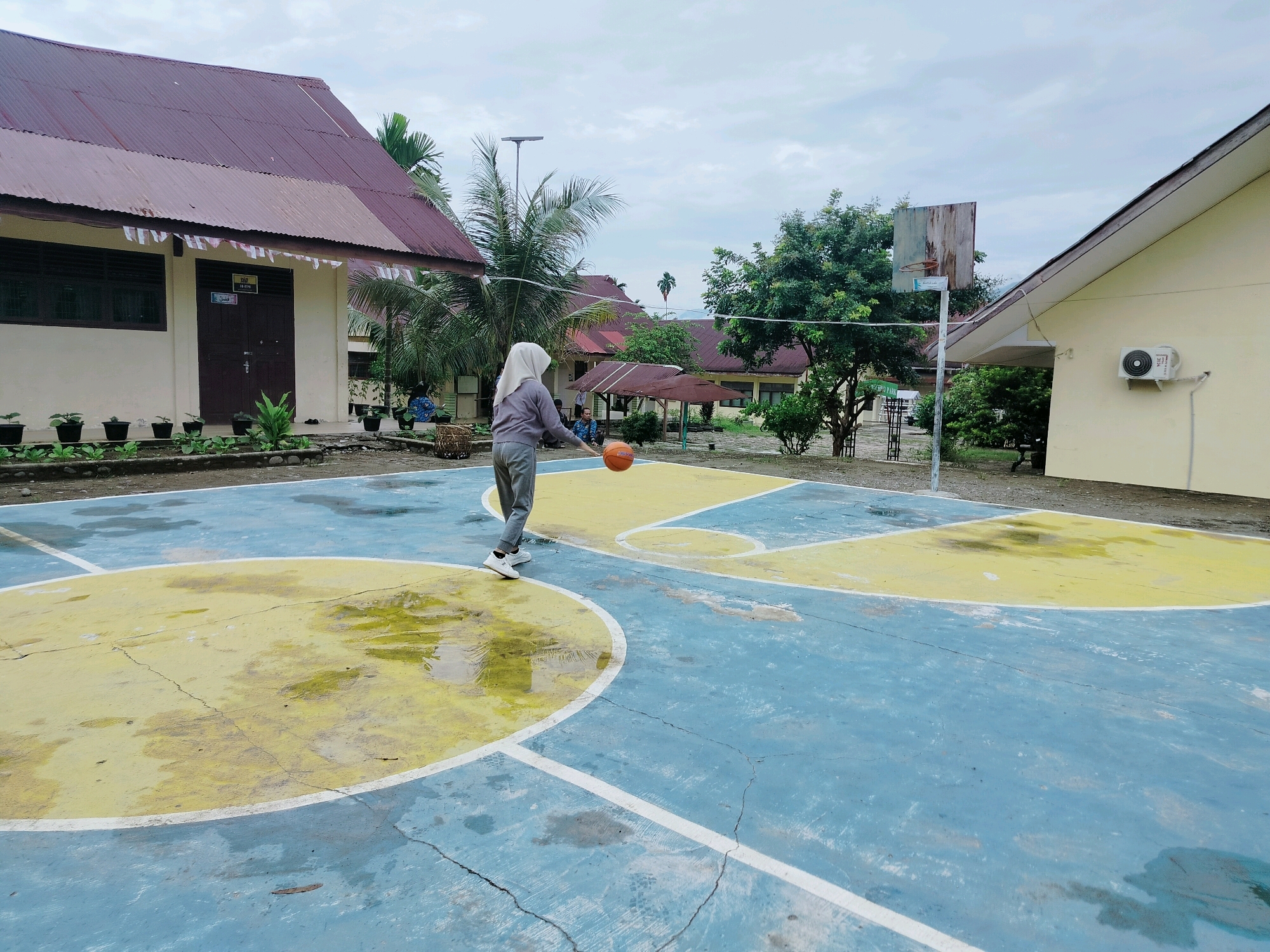 A young woman wearing a light grey hijab and athletic wear stands on a blue and yellow outdoor basketball court, holding an orange basketball. She is positioned in the middle of the court, facing towards the left side of the frame. The court appears wet in patches, suggesting recent rain. In the background, there are several buildings with red roofs, some of which appear to be school facilities, typical of a school compound. Trees and palm trees are visible around the buildings. A basketball hoop with a weathered backboard is situated on the right side of the court. The sky is overcast. The overall setting suggests a humid, tropical climate, consistent with Purwodadi, Indonesia.