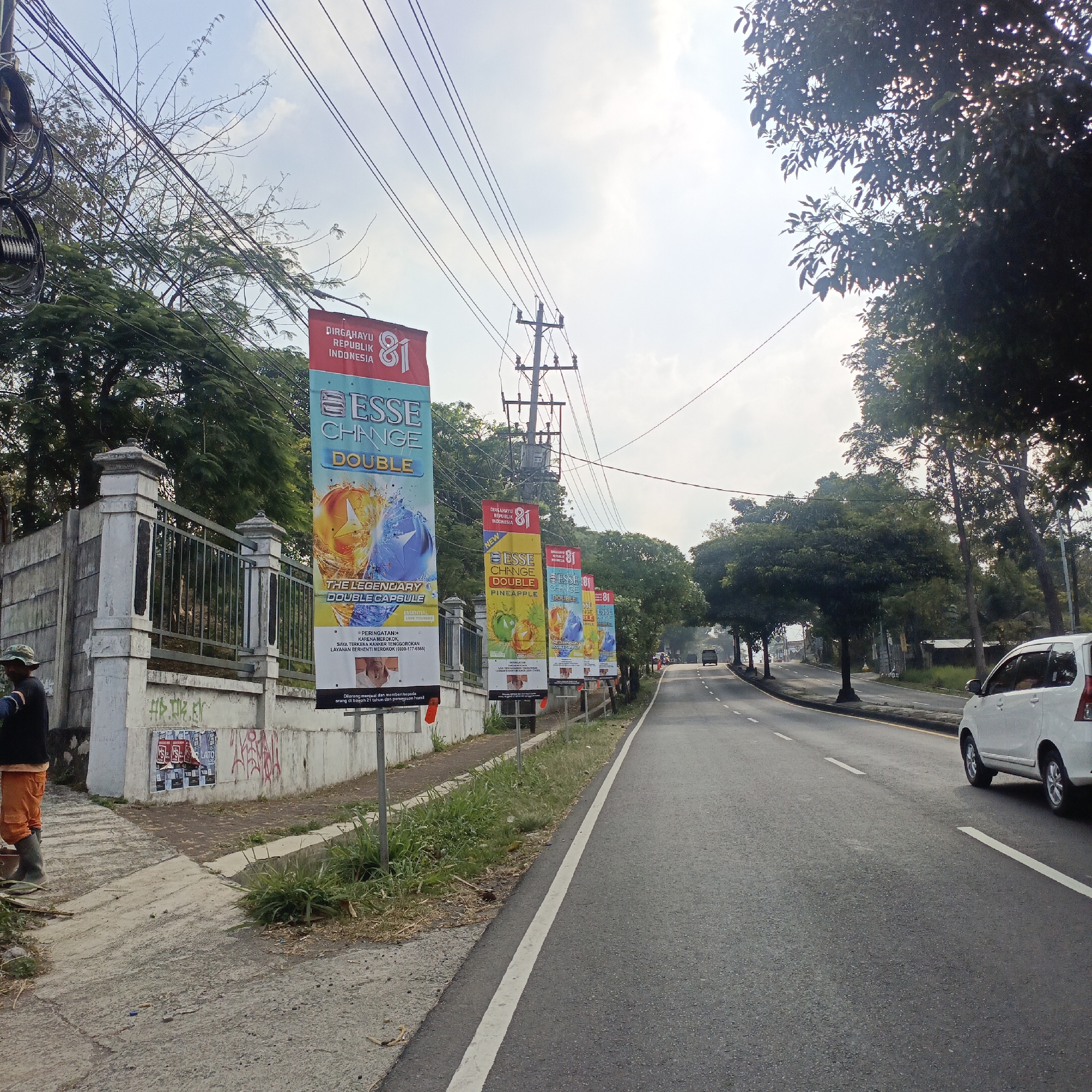 The image depicts a street scene in Salatiga, Indonesia, with a row of vertical banners advertising "ESSE CHANGE DOUBLE" products. The banners are placed along the sidewalk next to a road, with a white car visible driving on the right side. Electrical poles and wires stretch across the sky. On the left side of the image, a person wearing orange overalls and a hat is standing near a concrete wall with some graffiti. Trees line the street and the background, with a bright sky overhead. The banners are colorful and feature images of fruits and product packaging. One banner prominently displays the text "DIRGAHAYU REPUBLIK INDONESIA 81," indicating a national celebration. Another banner includes a warning about selling to individuals under 21 years of age and pregnant women.