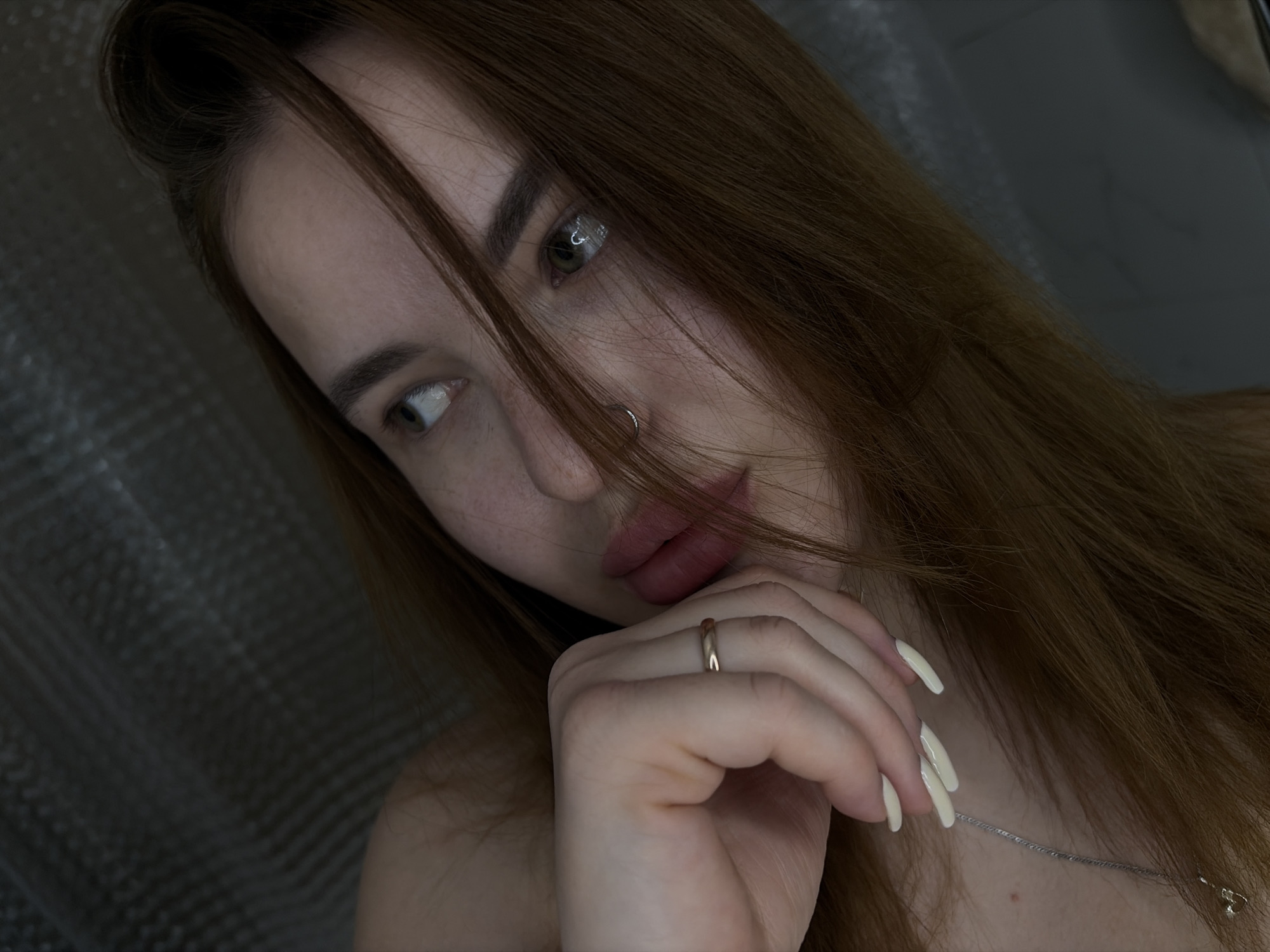 A young woman with reddish-brown hair and light skin is the primary subject of the image. She has a nose ring and is wearing a gold ring on her finger. Her hand is near her face, and she appears to be wearing a necklace. The background is out of focus, but a textured surface is visible. The image was not taken in Krasnodar, Russia.