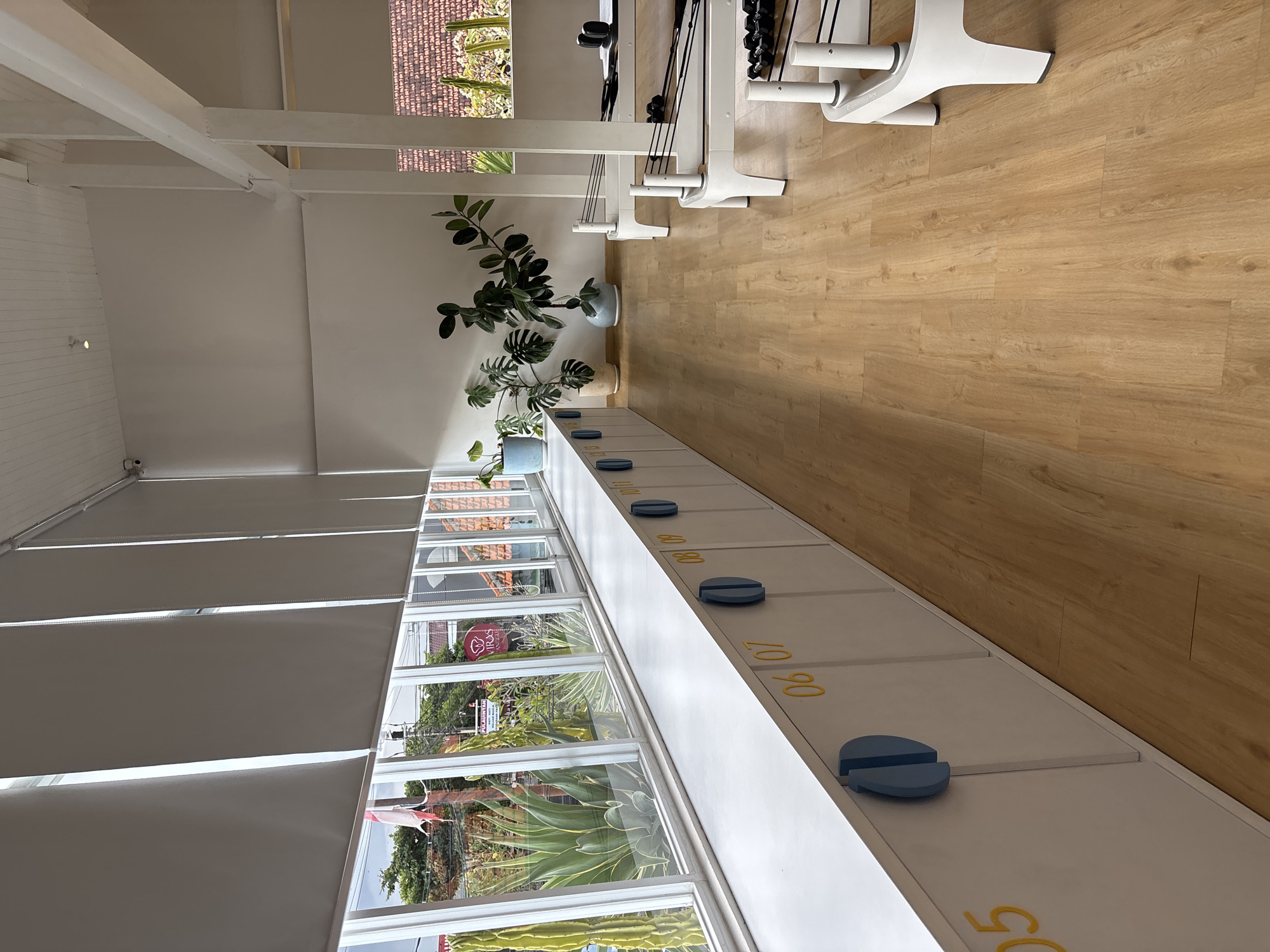 The image shows the interior of a building in North Kuta, Indonesia, identifiable by the view through the windows. The scene is bright with natural light. The room features a row of white lockers with blue circular handles, numbered in yellow. The window overlooks a street scene with greenery and some buildings. Additionally, there are indoor plants in pots near the lockers. Exercise equipment is visible in the background and a light-colored wooden floor.