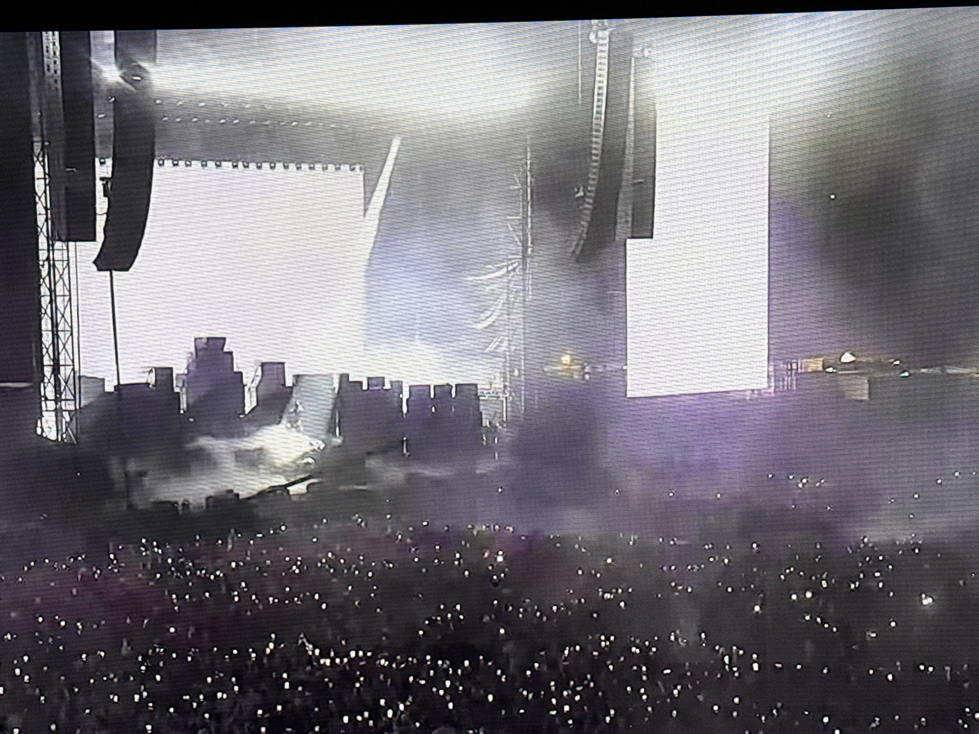 A large crowd of people is gathered at a concert or live performance. The crowd is holding up cell phones, their screens illuminating the dark scene. In the background is a stage set up with large screens and speaker arrays. There is smoke or haze in the air, with bright lights shining from the stage, creating a dynamic and energetic atmosphere. The location appears to be an outdoor venue. It is not possible to determine if this location is in the city of Sofia, Bulgaria from this image alone.