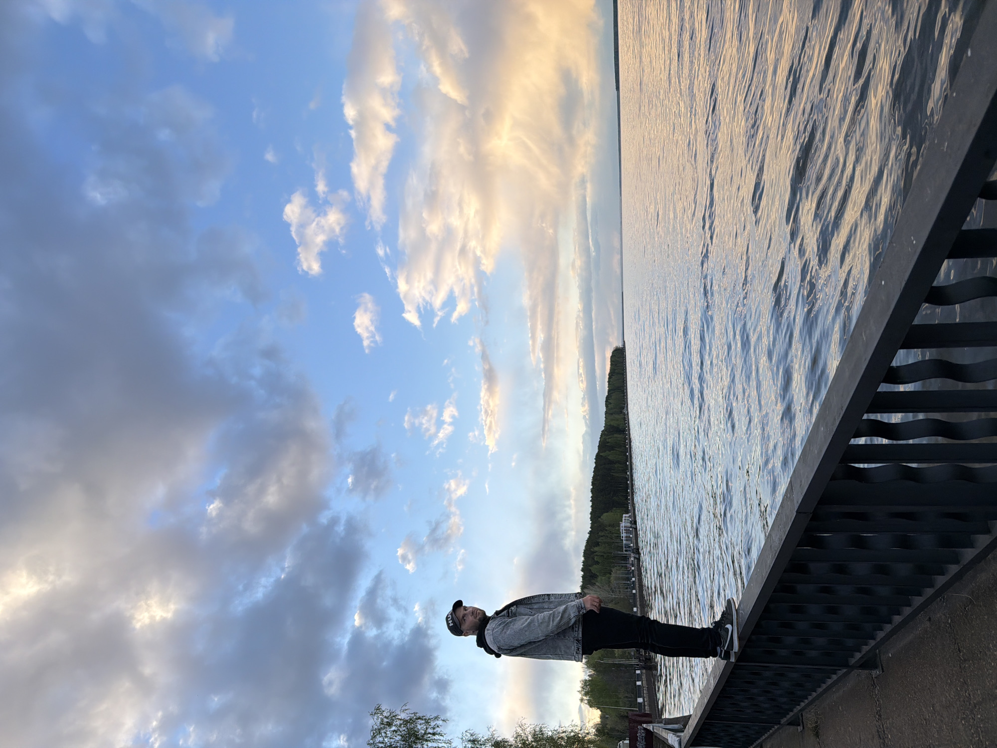 A person stands on a bridge railing, with their body oriented vertically against the horizontal view. The person is wearing a black cap, a grey denim jacket, and black pants with black and white sneakers. They are looking towards the camera.
Behind the person is a large body of water with a line of trees on the far shore. The sky is filled with clouds, displaying a mix of light and dark tones. The bridge railing is made of dark metal.
The location appears to be in the city of Chaikovsky, Russia, as indicated by the scene composition.