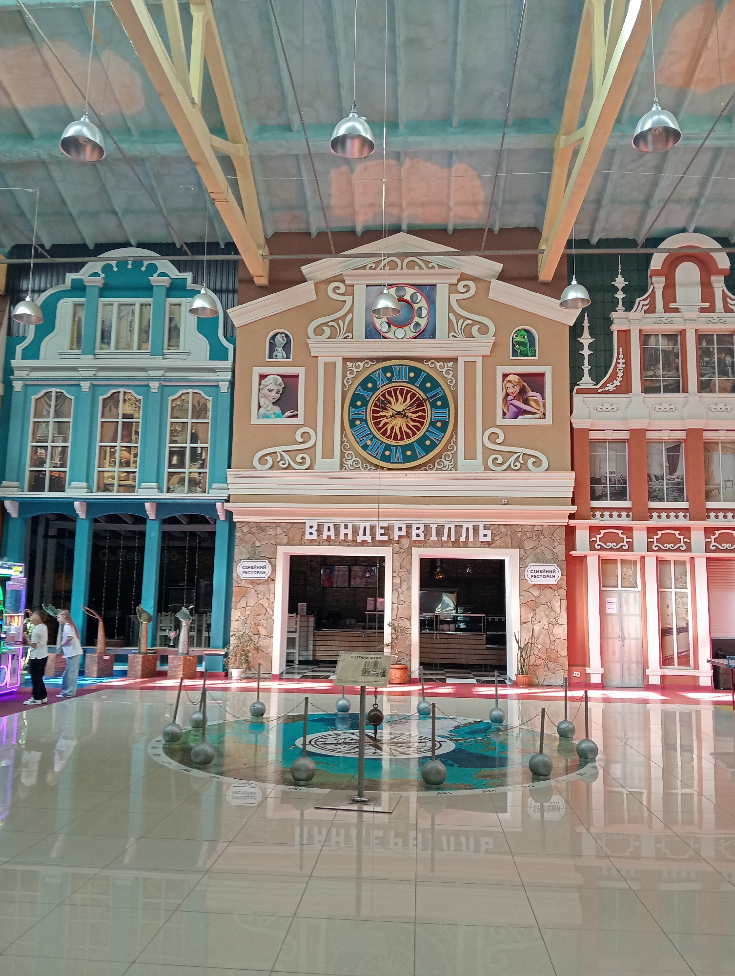 The image shows an indoor view in Lutsk, Ukraine. The scene features a brightly lit, polished floor with a circular installation containing a map design and several pendulum-like structures. Three building facades are centrally positioned in the background, designed to resemble Dutch architectural styles. The central facade features a large clock and portraits. Two women stand near the left side of the image. The lighting consists of metal dome-shaped pendant lights suspended from the ceiling.
