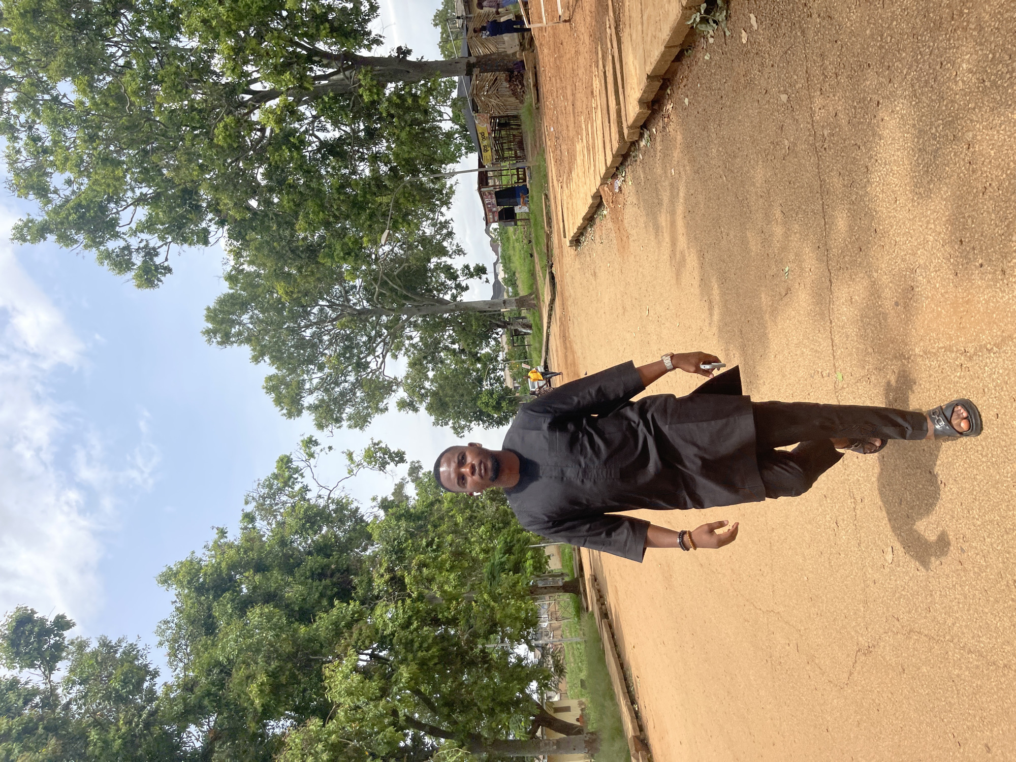 In Rafin Par, Nigeria, a man walks on a dirt road lined with trees. He is wearing a black traditional outfit and sandals, and he holds a phone in his right hand. The road is surrounded by green foliage and scattered buildings, with a few other people visible in the background. The sky is partly cloudy.
