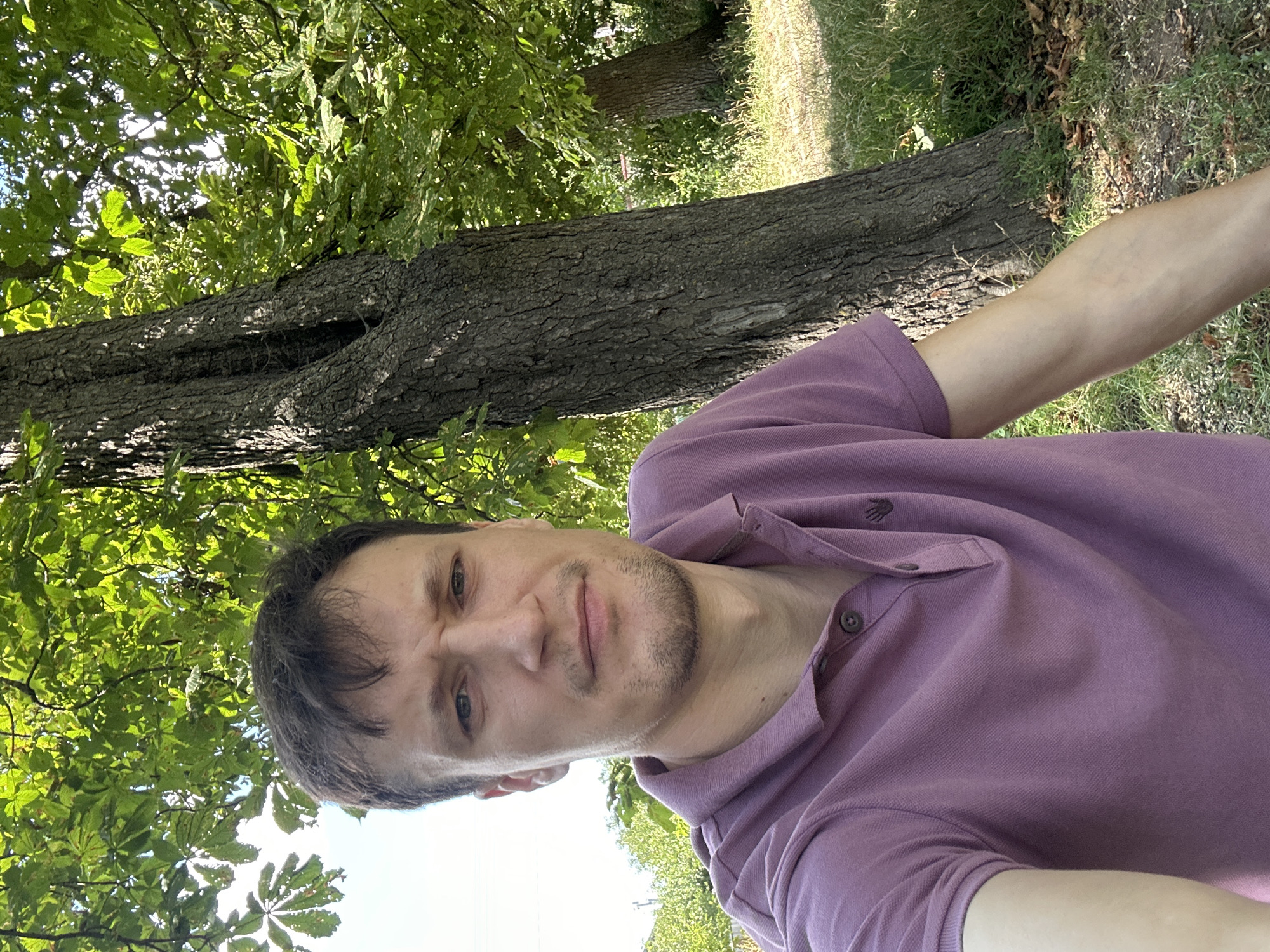 A close-up, waist-up selfie of a man with short brown hair and a light beard is taken outdoors. He is wearing a purple polo shirt and has a neutral expression. The man is positioned in front of a large tree with a rough, textured trunk and green leaves overhead. The background consists of more trees and grass, suggesting a park or natural setting. The lighting indicates it is daytime, with sunlight filtering through the leaves. The photo appears to be taken in Makiv, Ukraine.