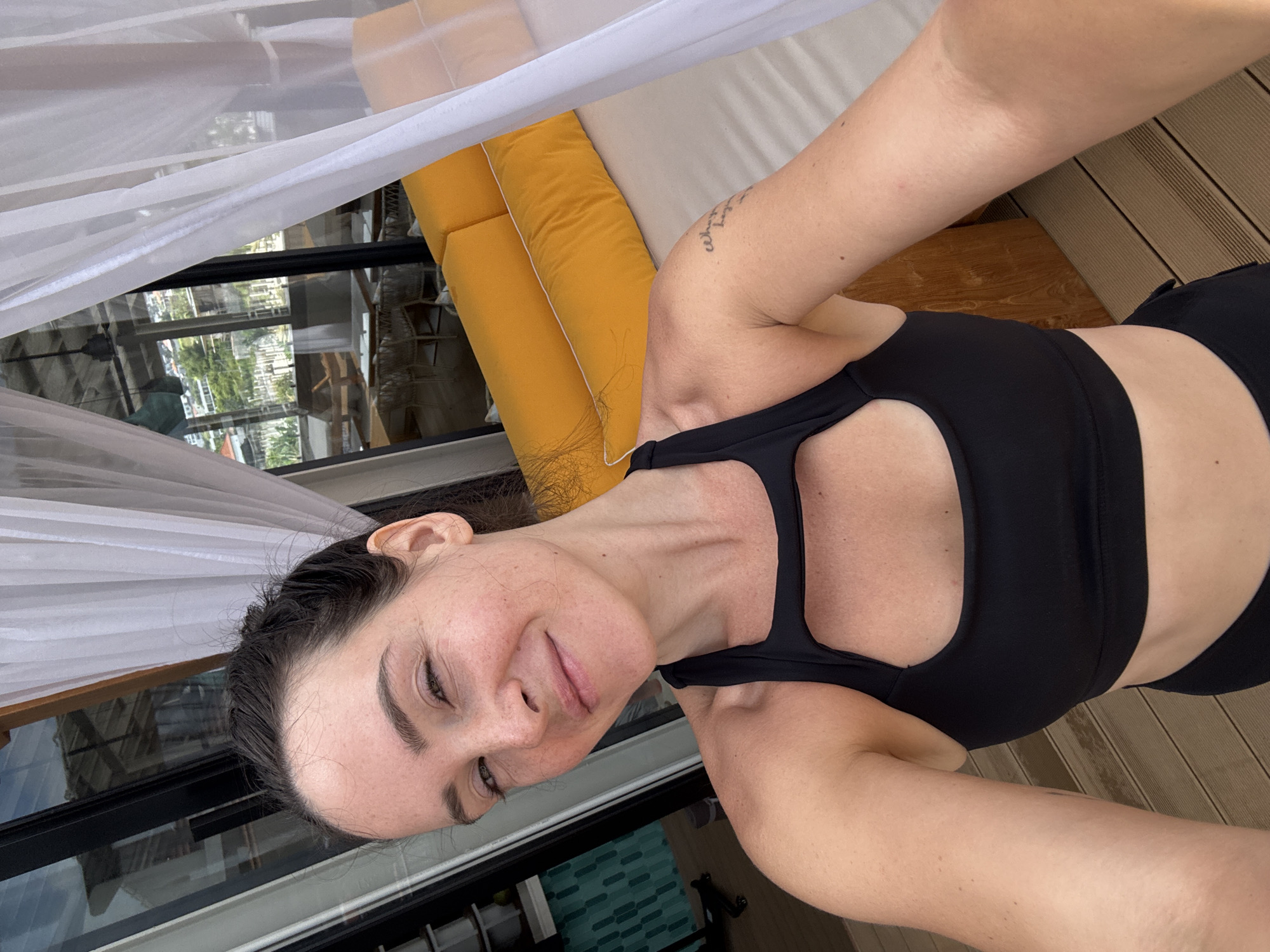A medium shot captures a woman with dark hair pulled back, taking a selfie outdoors. She's wearing a black sports bra and has freckles across her nose and cheeks. Behind her, white curtains billow, framing a view of a resort with yellow lounge cushions, wooden decks, and large windows reflecting a tropical setting. The background suggests a location in North Kuta, Indonesia, possibly a beach resort or villa.