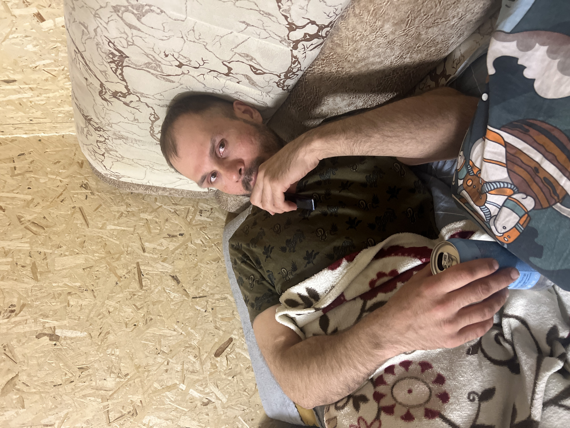 The photograph shows a man lying on a bed. He is in Chaikovsky, Russia. He wears a patterned t-shirt and holds a black object to his face in his left hand. He has a can in his right hand, resting on a patterned blanket. The bed has a patterned headboard, and a patterned blanket is partially covering him. The room has a wooden floor.