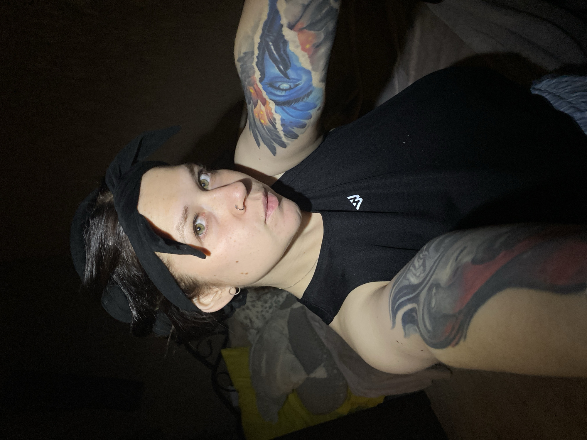 A selfie-style photograph depicts a light-skinned person with a black hair bow, visible tattoos on both upper arms, and a nose ring. The person is wearing a black top. The background is mostly dark and indistinct, suggesting a poorly lit indoor location. There is no information to determine if this picture was taken in the city of Chaikovsky, Russia.
