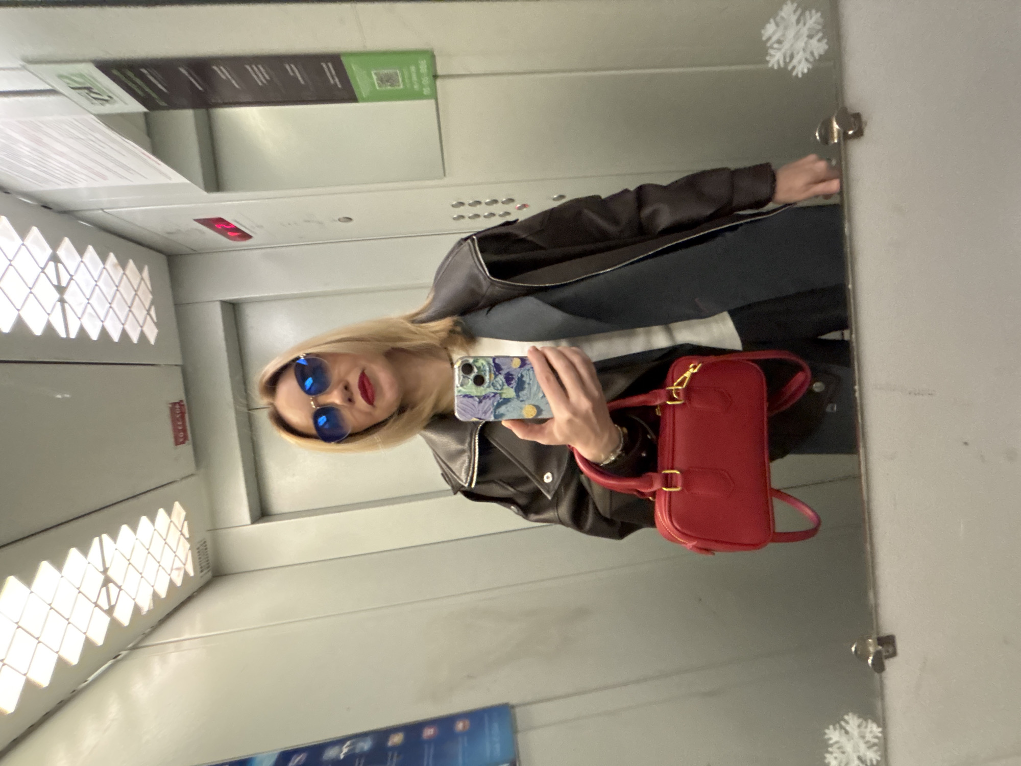 A person with blonde hair, wearing blue sunglasses and a black leather jacket, is taking a selfie in an elevator. The person is holding a smartphone with a patterned case in their left hand. A red purse hangs from their shoulder. The elevator interior has a metal finish, and there are various panels, vents, and control buttons visible. A snowflake sticker is visible on the mirrored wall of the elevator. There's also some type of advertisement or notice, but it's not clearly visible. The location of the elevator is Unknown, Unknown.