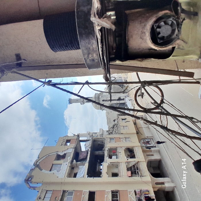 The image captures a scene of extensive urban damage in Khan Yunis, Palestinian Territory, from an elevated vantage point. In the immediate foreground on the right, a makeshift metallic apparatus features a dark cylindrical object stacked on a larger unit with an exposed fan, interconnected by multiple wires and cables extending across the frame. To the left, a multi-story building, light-colored with decorative archways, exhibits severe structural damage on its upper floors, with exposed interiors and missing sections of walls; laundry is visible hanging from balconies on the lower levels. A single individual, dressed in dark attire, walks on the paved street below, moving away from the viewer. The background is dominated by the heavily damaged Great Mosque of Khan Yunis, identifiable by its partially intact minaret and remnants of its dome, surrounded by widespread rubble and debris from destroyed structures. The sky above is partly cloudy.