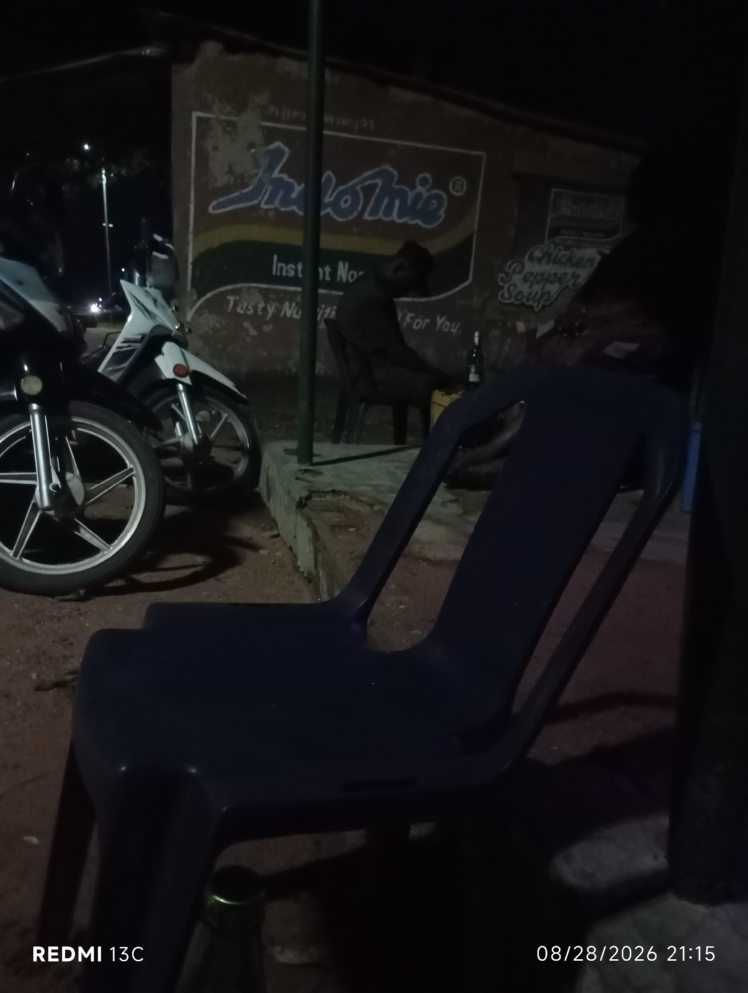 In Shendam, Nigeria, at night, a dimly lit scene unfolds outdoors with a focus on a dark blue plastic chair in the foreground. Behind it, two motorcycles are parked. The wall behind the motorcycles features a faded advertisement with the prominent "Indomie" logo, alongside text that appears to say "Instant N... Tasty Nu... For You." to the left, and "Chicken Pepper Soup" to the right. A person is seated on a chair to the right of the frame, facing away from the camera. The ground appears to be dirt or gravel, and a bottle with a green cap is visible near the bottom of the frame. The lower right corner displays a timestamp: "08/28/2026 21:15" and "REDMI 13C".