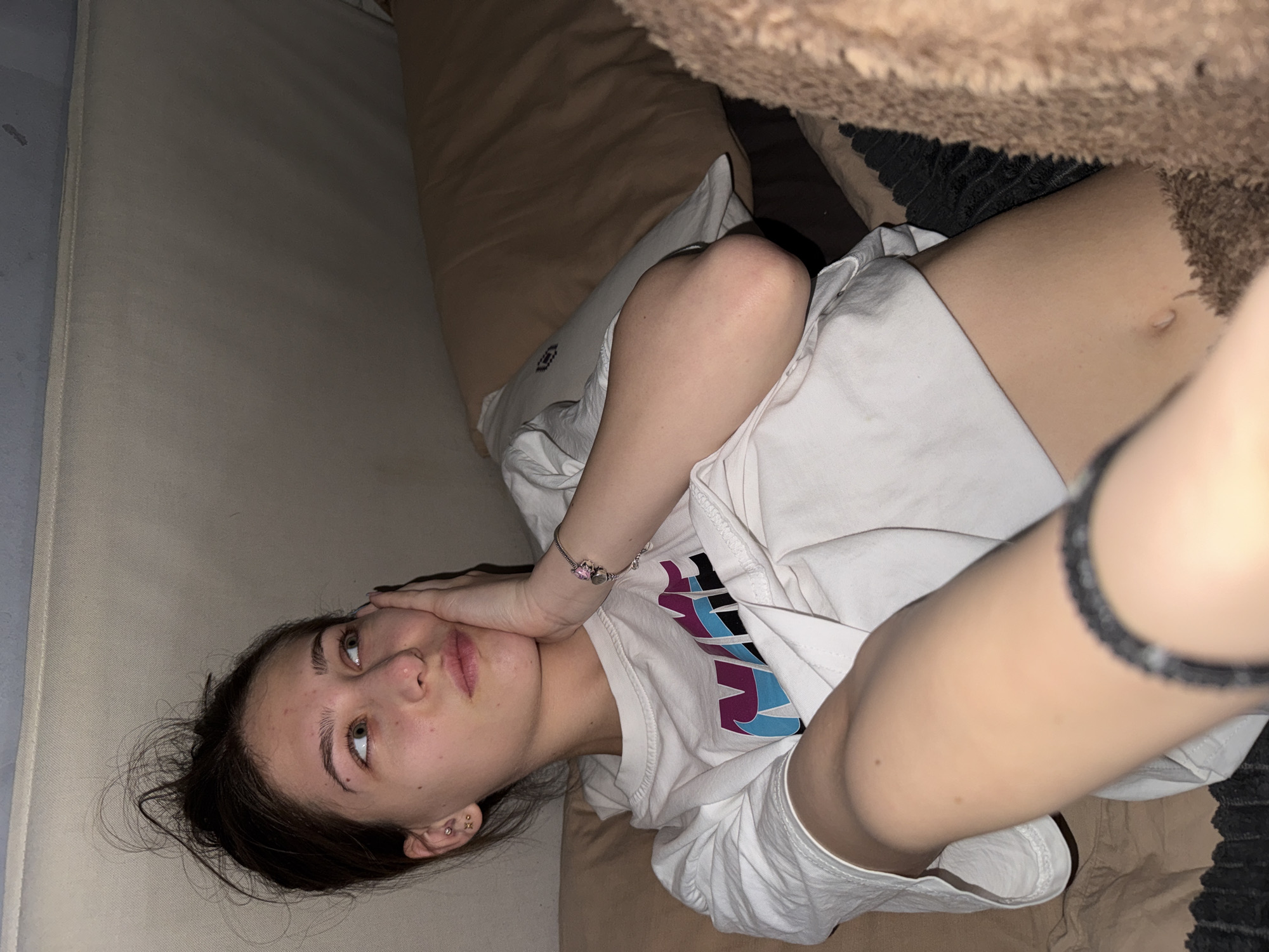 A young woman lies on a beige upholstered surface with a large tan pillow behind her head and shoulders, and a brown, fuzzy object, possibly a stuffed animal or blanket, in the upper right corner of the frame. She is wearing a white t-shirt with a design featuring purple, blue, and black colors, and possibly shorts. Her right hand is touching her face. The background suggests an indoor environment with a light source illuminating the scene. The location is not discernible from the image.