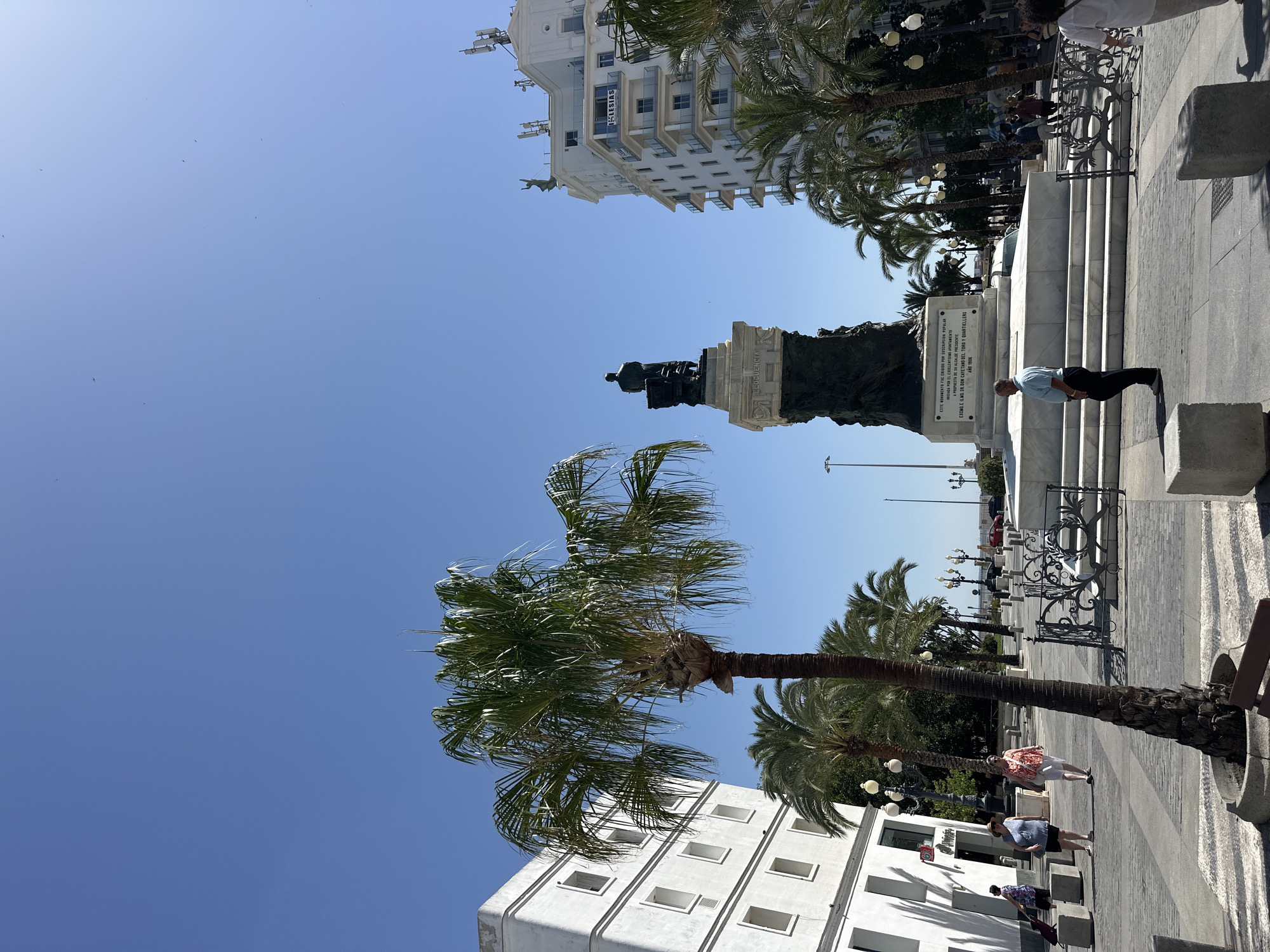 The image depicts a daytime scene in a public square in Cádiz, Spain, likely the Plaza de las Cortes. In the center of the square stands a large monument with a statue of a seated figure atop a substantial stone base. The base features an inscription, although the text is not fully legible. Palm trees are scattered throughout the square, with one prominent palm tree in the foreground to the left.
Several people are visible in the image. A man in a light blue shirt and dark pants stands near the monument, facing away from the camera. To the left, a group of women are walking, one pushing a shopping cart. In the background, more people are seen strolling through the square, and some are near buildings.
The environment includes paved walkways with patterned sections, decorative metal fencing around the monument's base, and lampposts. White buildings with multiple windows line the sides of the square. In the background, taller white buildings with balconies and visible cell towers stand against a clear blue sky. The overall impression is of a sunny day in a lively urban setting.