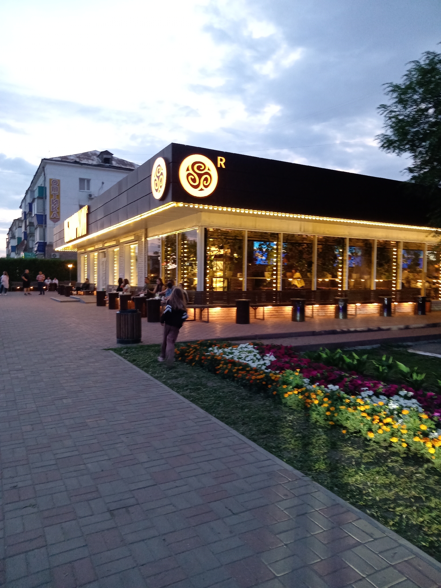 The image shows an outdoor scene in the city of Rudny, Kazakhstan, likely in the evening, with a restaurant or cafe illuminated by string lights. The building has a dark facade with large glass windows and a prominent sign featuring a circular emblem and the letter "R". In front of the building, there are tables and chairs set up outdoors, and several people are seated and standing. A brick pathway leads away from the building towards the foreground, bordered by a flowerbed with a variety of colorful flowers. To the left of the building, another building with windows and balconies is visible, and a sign reading "ОПТИКА" (Optika - Optics) is affixed vertically to its facade. A few people are walking on the pathway. The sky is overcast, and the lighting suggests dusk or early evening.