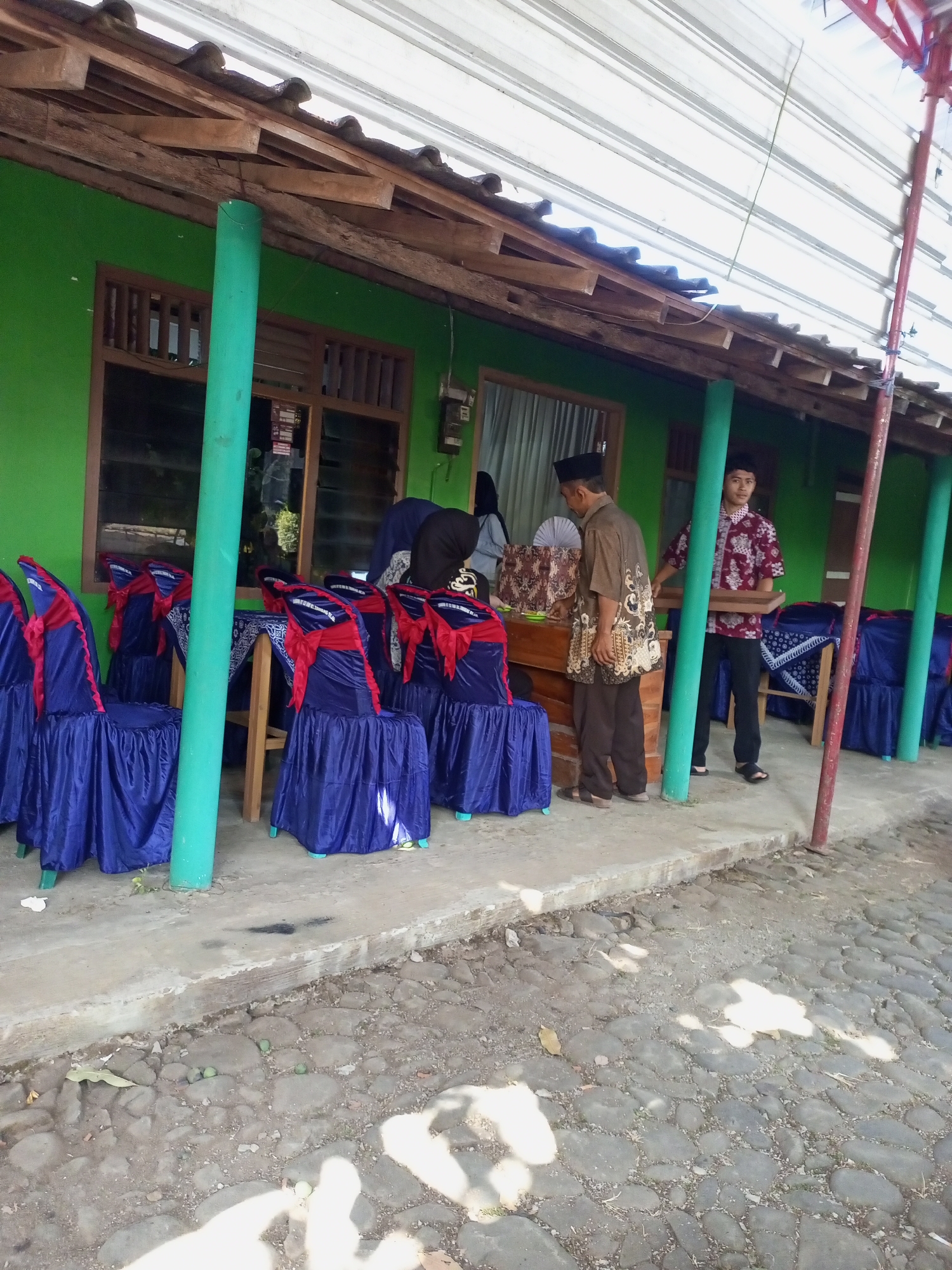 The image depicts an outdoor event setup in Jatisrono, Indonesia, likely a gathering or ceremony. In the foreground, several chairs are draped in blue fabric with red sashes tied around their backs, suggesting a formal or festive occasion. A concrete pathway borders the area, which transitions into a cobblestone ground. The background features a building with a bright green painted wall and wooden window frames, covered by a sloping roof constructed with wooden beams and corrugated metal sheeting. Several people are present, including two men in traditional Indonesian attire. One man, wearing a batik shirt and a peci (cap), appears to be interacting with a table set up with items. Another younger man in a patterned red shirt and black pants is carrying a wooden tray. Other individuals, partially obscured, are also visible near the building. The overall scene suggests a community event taking place in a residential or public outdoor space in Jatisrono.