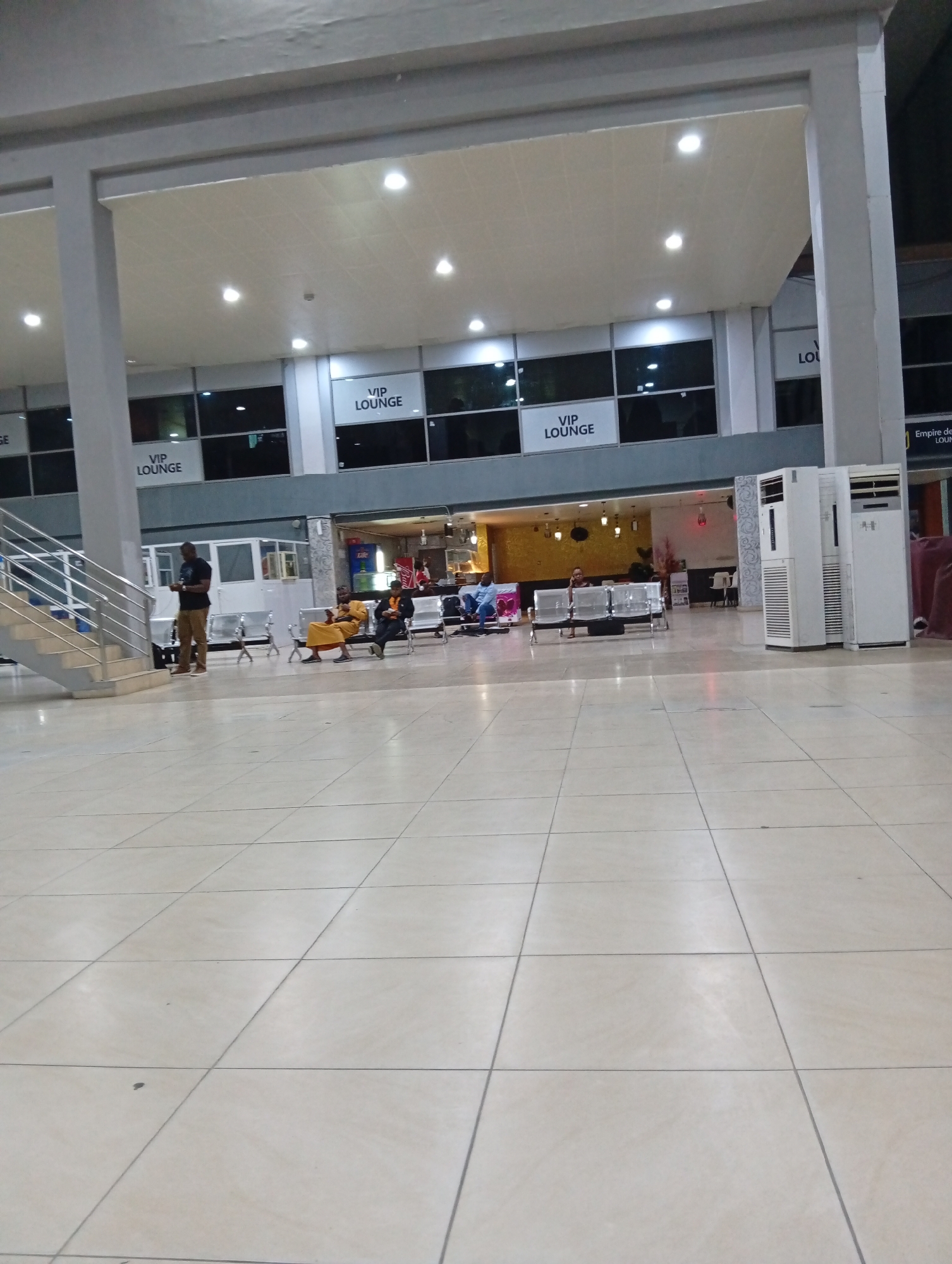 The image shows the departure lounge of Port Harcourt International Airport in Omagwa, Nigeria. The lounge is an open area with high ceilings, tiled flooring, and large windows labeled "VIP LOUNGE". People are seated on metal benches, and a man in a dark t-shirt and khaki pants is standing near a staircase, looking at his phone. Several portable air conditioning units are positioned near the edge of the open area. The scene appears calm, with a mix of individuals waiting.