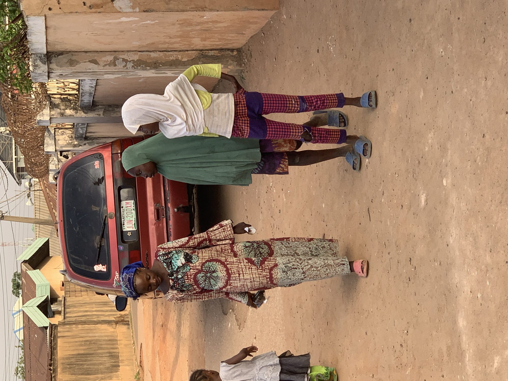 The image captures a scene in Jos, Nigeria. Three women stand near a red minivan with the license plate "JJN 200XM" from the Plateau State. Two girls, wearing head coverings and dressed in casual wear, stand together beside the van. An older woman in a patterned dress and blue head wrap is nearby, holding a small object. The ground appears to be dirt, and there are buildings in the background.