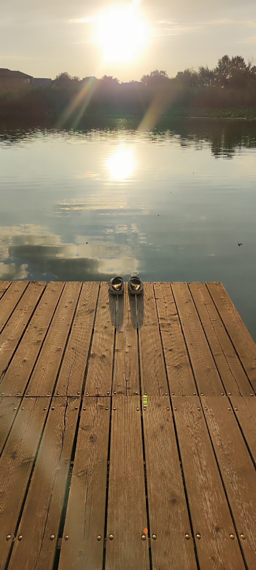 A pair of dark, rubber slip-on shoes are placed side-by-side on a wooden pier that extends out over a body of calm water. The pier is constructed of wooden planks running lengthwise, with visible nails securing them. The sun is low in the sky, casting a warm, golden light and creating a bright glare on the water's surface. The glare also causes lens flare on the sun's rays in the upper portion of the image. The water reflects the sky, clouds, and distant trees and buildings. The opposite shore of the water is lined with trees and some structures, suggesting a rural or suburban setting. The scene appears to be located near Starokorsunskaya, Russia, given the context of the query. The overall impression is one of tranquility and the end of a day.