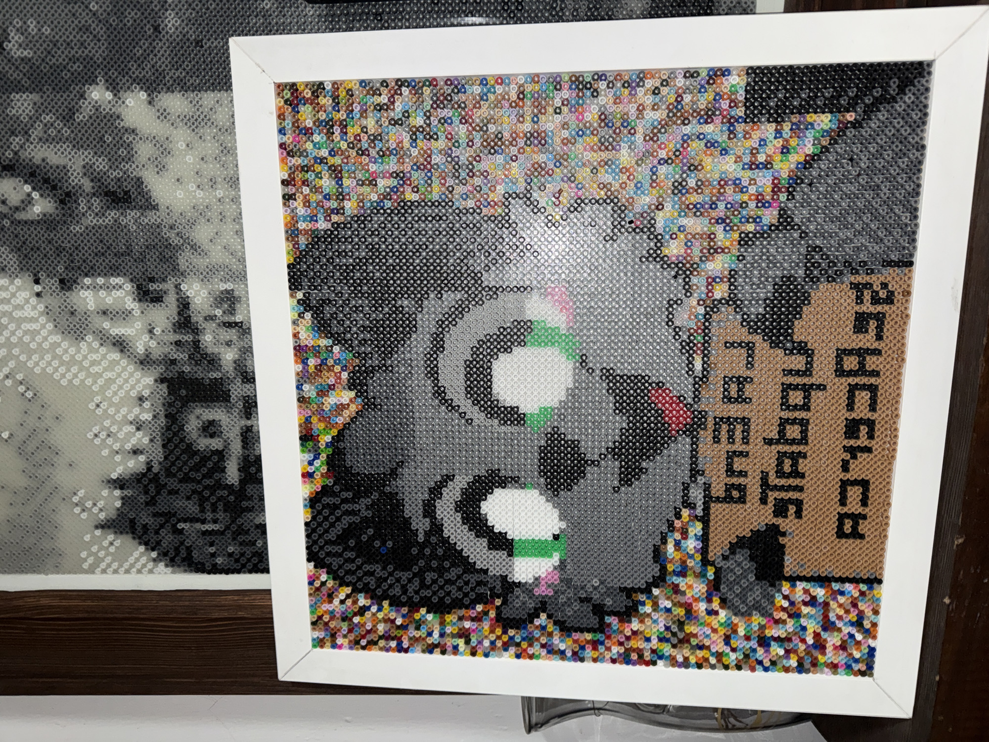 The image displays two pixel art renderings in frames. The one on the right, with a white frame, appears to be a stylized depiction of a raccoon holding a sign, created with Perler beads. The sign reads "Trabajo por tacos", written in vertical, brown-colored beads. The surrounding area is filled with multicolored beads, giving a textured background. The picture on the left, partially visible with a wooden frame, appears to be a grayscale image, also rendered in Perler beads. The environment suggests an indoor setting, perhaps an art display or studio. The location of Puebla, Mexico, is inferred solely from the Spanish phrase on the sign.