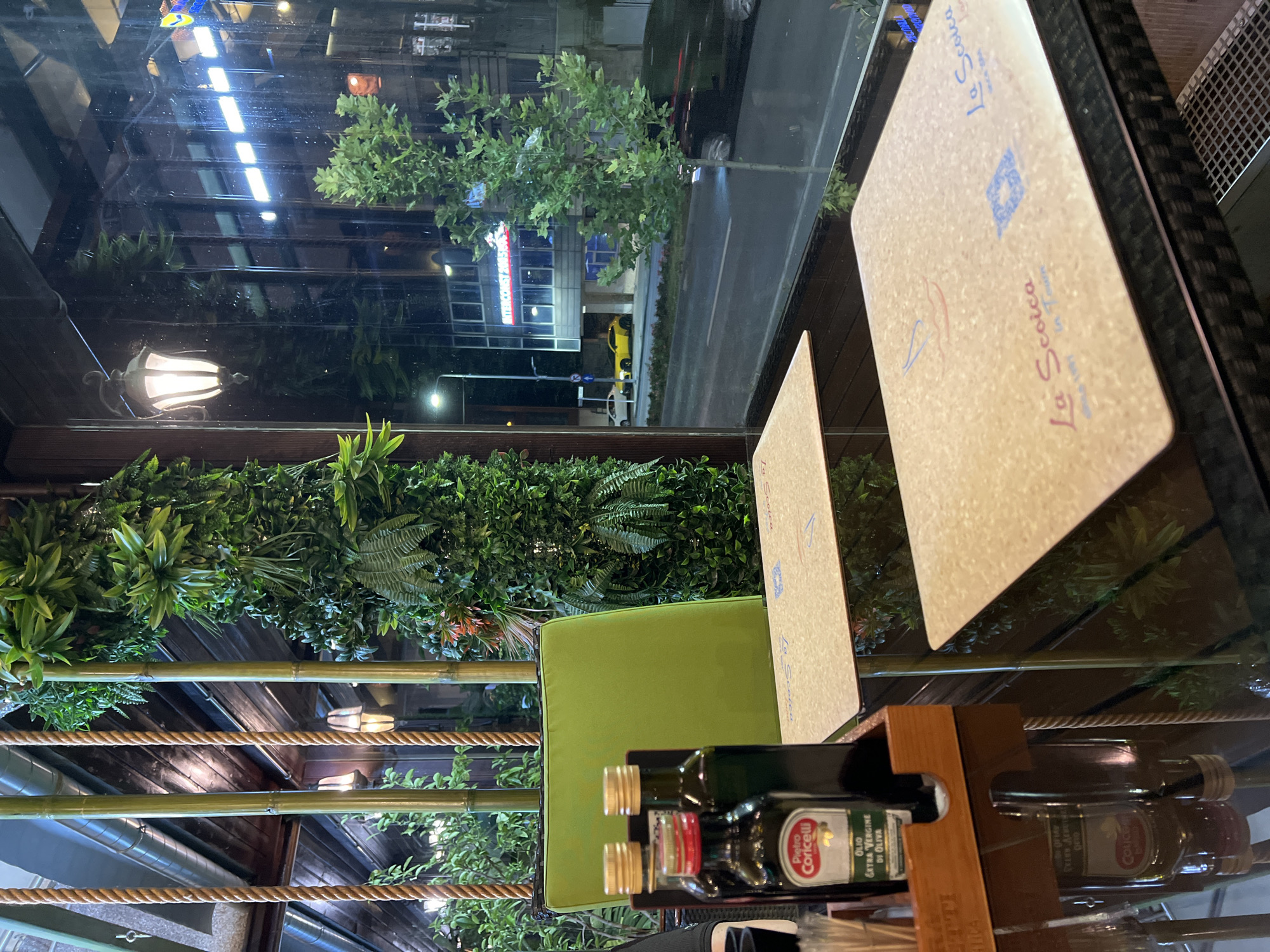 The image depicts a table setting at an outdoor seating area of a restaurant, likely located in Constanța, Romania, given the architectural style and the presence of a prominent building with neon signage visible through the window. The table is made of dark glass and is set with two placemats, each featuring the restaurant's logo, "La Scoica." On the table, there are two bottles of Pietro Coricelli Extra Virgin Olive Oil, one with a red label and the other with a green label. To the left of the olive oil bottles, a wooden holder contains cutlery. The background features a wall covered with artificial greenery, bamboo stalks, and hanging ropes. Through the window, a street scene is visible at night, with streetlights, cars, and a building with illuminated signage. A bright yellow car is parked on the side of the street. The overall ambiance suggests a dimly lit, naturalistic setting within an urban environment.