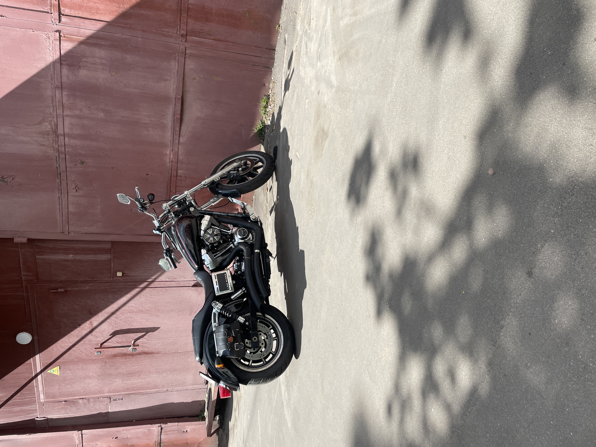 A black motorcycle is parked on an asphalt surface next to a reddish-brown metal wall. The wall is constructed of rectangular panels with vertical seams. A shadow of foliage is cast onto the asphalt surface. The location is suggested as Khimki, Russia.