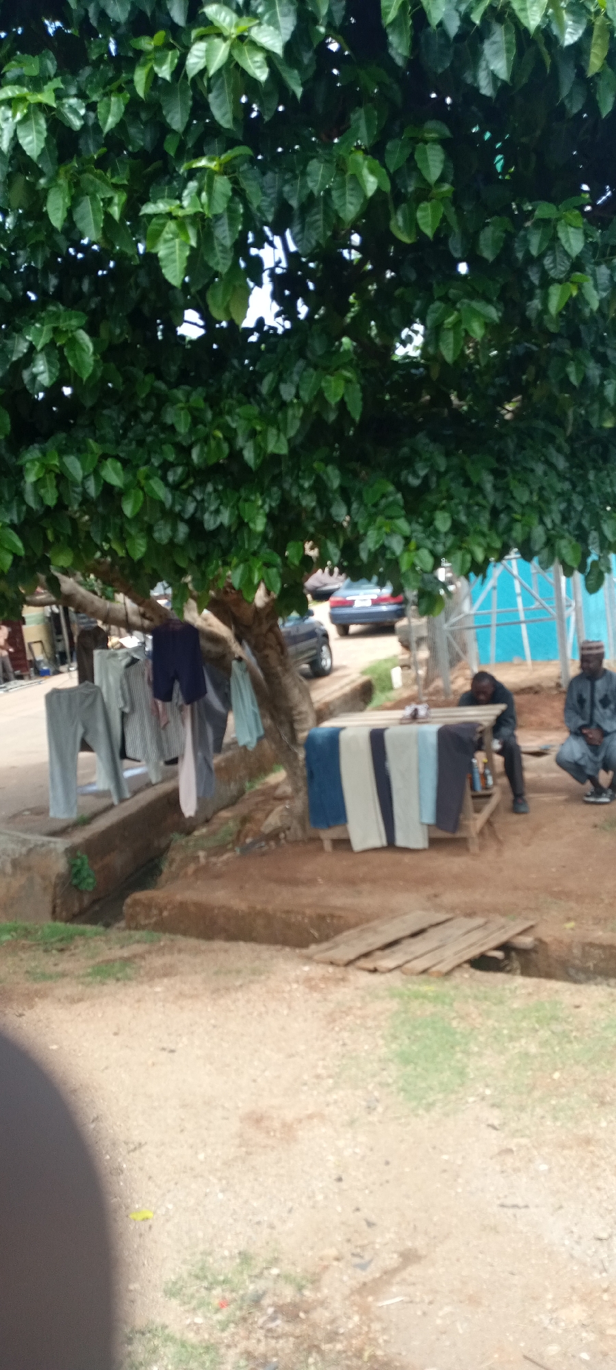 The image shows an outdoor scene in the city of Unknown, Unknown. A large tree with dense green foliage dominates the upper portion of the frame. Beneath the tree, on a raised platform, clothes are hanging to dry. A wooden table is laden with more articles of clothing. Two men are present; one sits at the table, and the other is squatting near the table. In the background, a car is parked on the street. A makeshift bridge of wooden planks spans a ditch or shallow trench in the foreground. The ground is predominantly dirt and gravel.