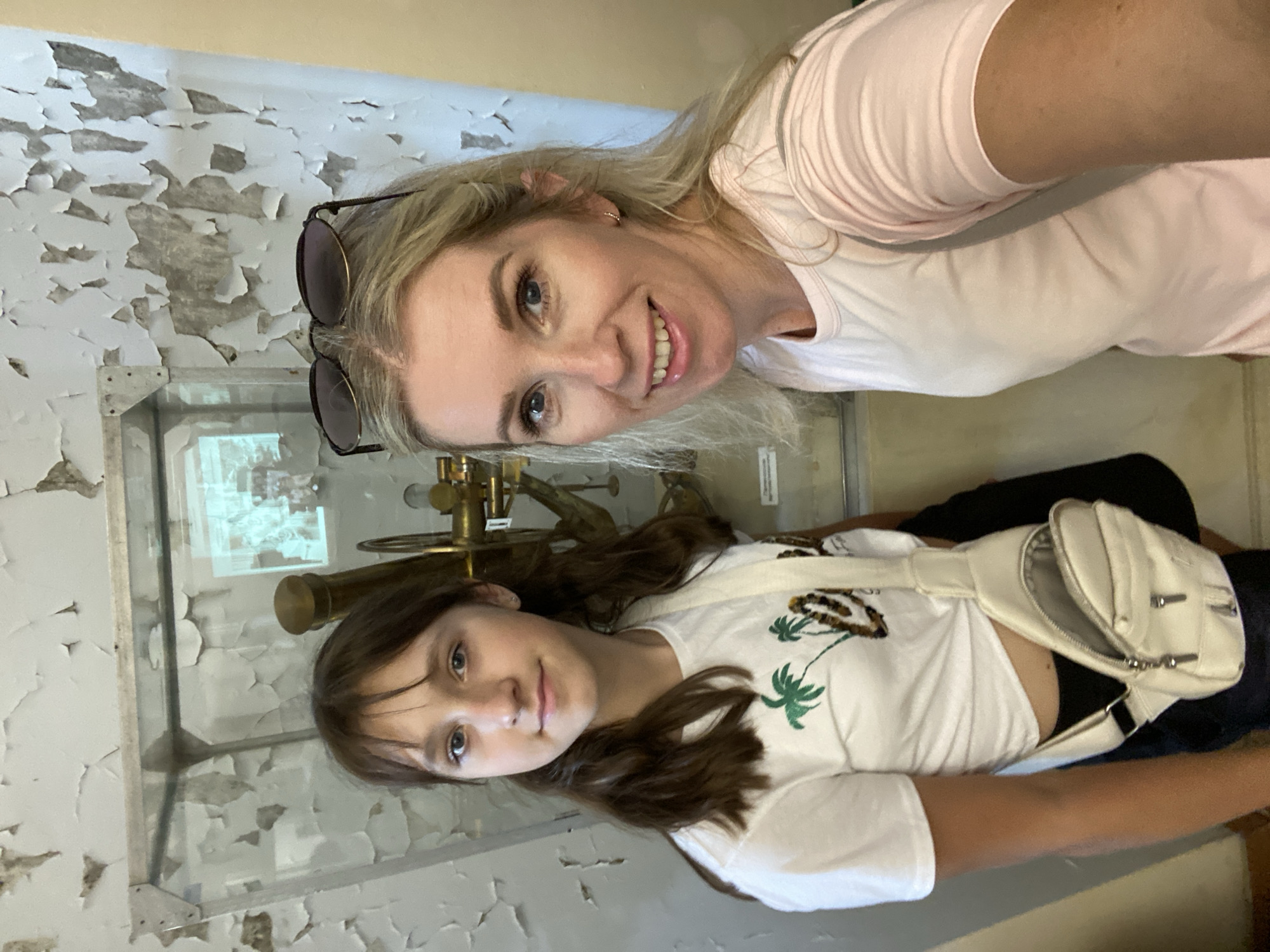 In a close-up, selfie-style photograph taken indoors, two females, likely mother and daughter, pose for the camera. The younger female, on the left, has shoulder-length brown hair and is wearing a white t-shirt with a palm tree graphic. She has a white fanny pack worn across her chest. The older female, on the right, has blonde hair and is wearing a light pink t-shirt. She has sunglasses perched on top of her head. They are standing in front of a glass display case containing what appears to be an old telescope. The background wall is textured with peeling paint, suggesting an aged or possibly neglected interior. The lighting is natural and appears to be coming from the front.