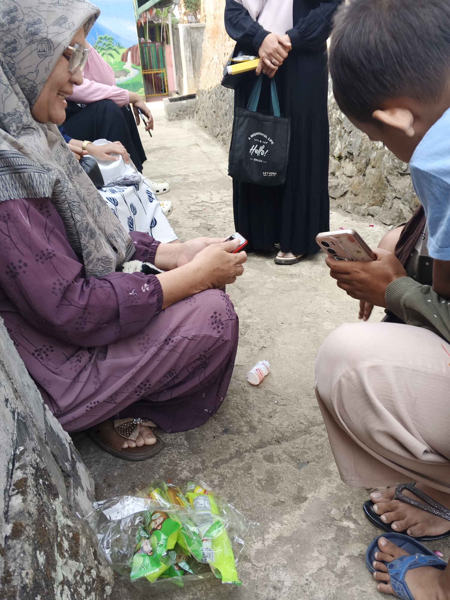 The image depicts a group of people outdoors in Warungkiara, Indonesia. In the foreground, a woman wearing a purple patterned dress and a headscarf is seated, looking at her phone. Next to her, a child in a light blue shirt is also looking at a phone. A small plastic bottle is on the ground near them. Further back, another woman in a black dress stands, holding a small tote bag and a yellow object. The background shows a wall and some colorful structures, suggesting an outdoor recreational area or a market. A bag of green-colored snacks is visible in the lower left corner.