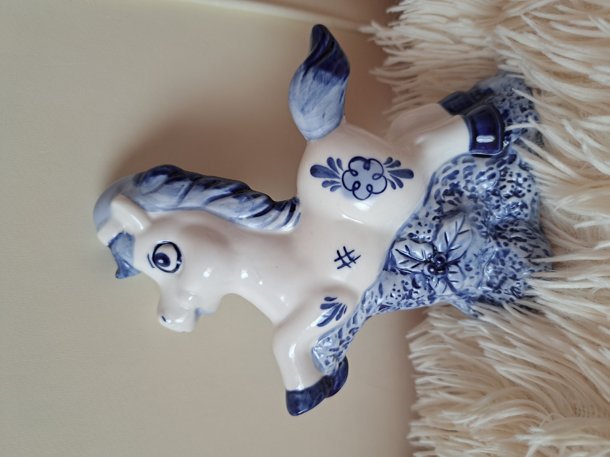 A white ceramic horse figurine with blue floral and decorative patterns is positioned on a fluffy white surface, resembling faux fur. The figurine has a stylized mane and tail, and its body is adorned with intricate blue designs. The horse is depicted in a playful pose, with its front legs raised. The background is a plain, light-colored surface. No specific location in Irkutsk, Russia can be identified.