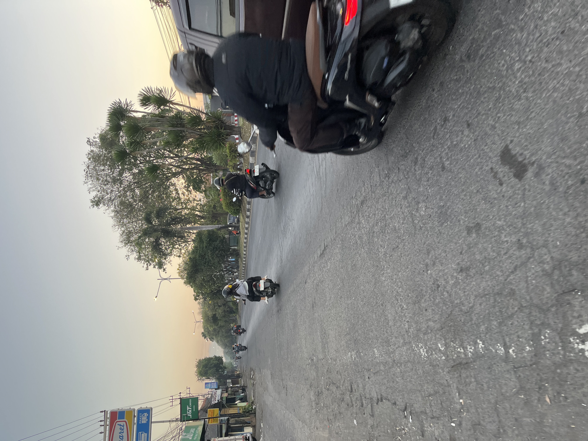A street scene at dusk in Ngaban, Indonesia, captures the hazy atmosphere with a clear sky transitioning from pale blue to a soft orange near the horizon. The road, made of asphalt, shows signs of wear and tear with visible cracks and patches. Several motorbikes are in motion. In the foreground, a person is riding a dark-colored motorbike, their back to the camera, wearing a black jacket and brown pants, and a dark helmet. To the left and further down the road, another person on a motorbike, also wearing a dark helmet, is seen from behind. Further ahead, more motorbikes are visible, some with riders. On the left side of the road, shops and businesses line the street, with prominent signs for "Indomaret," "J&T CARGO," and "DAGING AYAM." Streetlights are positioned along the road. Trees and foliage are scattered along the roadside, with taller, palm-like trees and other leafy varieties. A dark-colored van is parked on the right side of the road, partially obscuring the view of the motorbike in the foreground. The overall impression is a typical evening commute in a bustling Indonesian town.