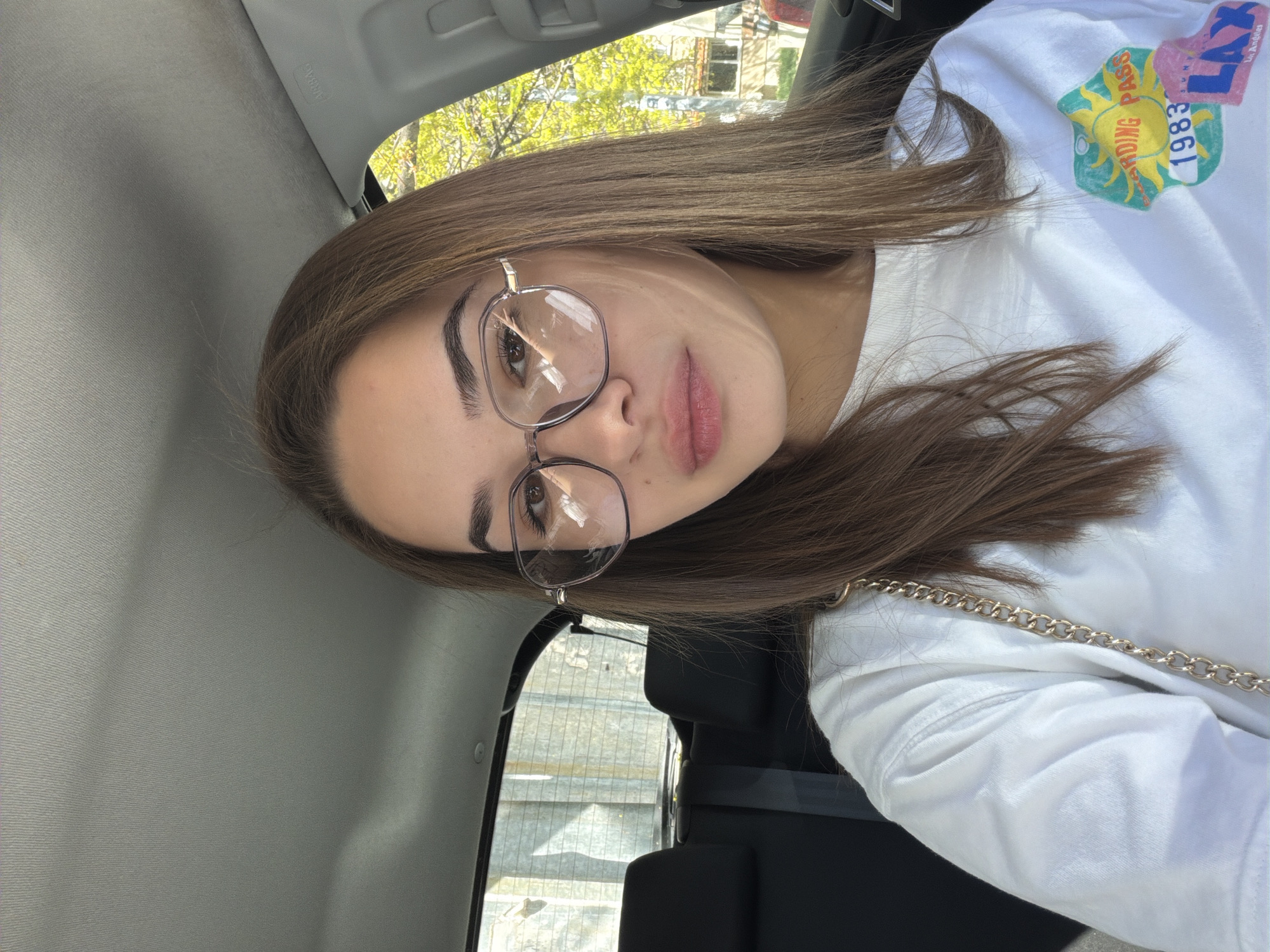A young woman with light brown hair, wearing glasses and a white t-shirt with a colorful graphic print, is sitting inside a car. The interior of the car is visible, including the ceiling, door panel, and seat. Outside the car window, buildings and trees are visible, potentially indicating an urban environment. Given the prompt's request, the location could be Sofia, Bulgaria, though specific landmarks are not discernible in the image to confirm.