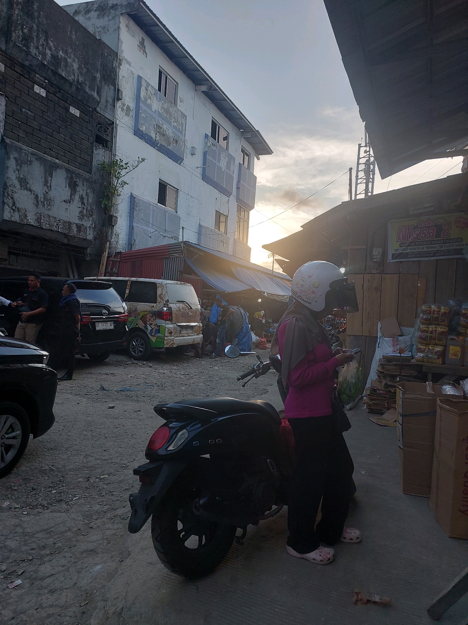 In Ambon, Indonesia, a street scene unfolds with a mix of vehicles and buildings under a hazy sky. A woman wearing a white helmet and pink shirt stands near a black motorcycle, holding a phone and a plastic bag. Two SUVs are parked nearby; one is black, and the other is decorated with cartoon characters. In the background, a multi-story building with weathered blue and white paint stands next to a more dilapidated structure. A small shop with goods displayed on shelves is visible on the right, with a sign for "Toko Nissa 2." The ground is a mixture of dirt and gravel, with debris scattered around. The overall atmosphere suggests a commercial area during the late afternoon, with the sun casting long shadows.