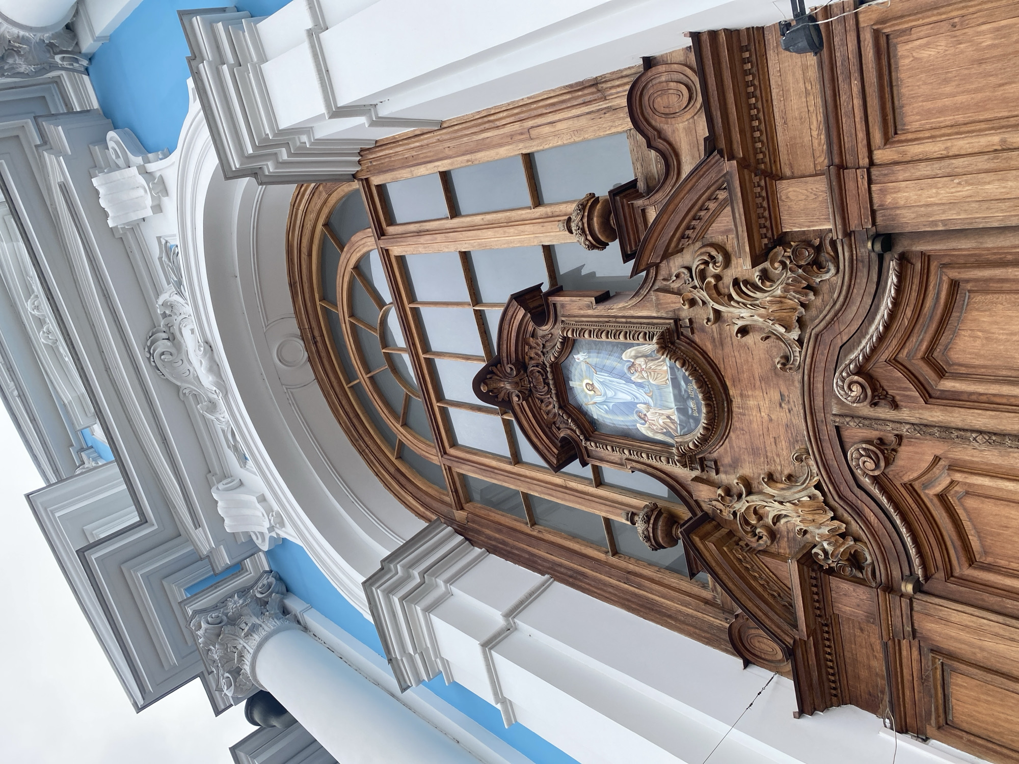This is a low-angle, close-up exterior shot of the ornate wooden doors and arched window of the Smolny Cathedral in Saint Petersburg, Russia. The doors are made of dark, polished wood with intricate carvings, including decorative scrollwork and architectural moldings. Above the doors is a large, arched window with multiple panes, also framed in wood. Centered above the doors, set into a decorative wooden surround, is a framed religious icon depicting Jesus Christ and several angels. The building's facade features white neoclassical architectural elements, including columns and detailed stucco work, contrasting with the bright blue exterior walls. The sky above is overcast.