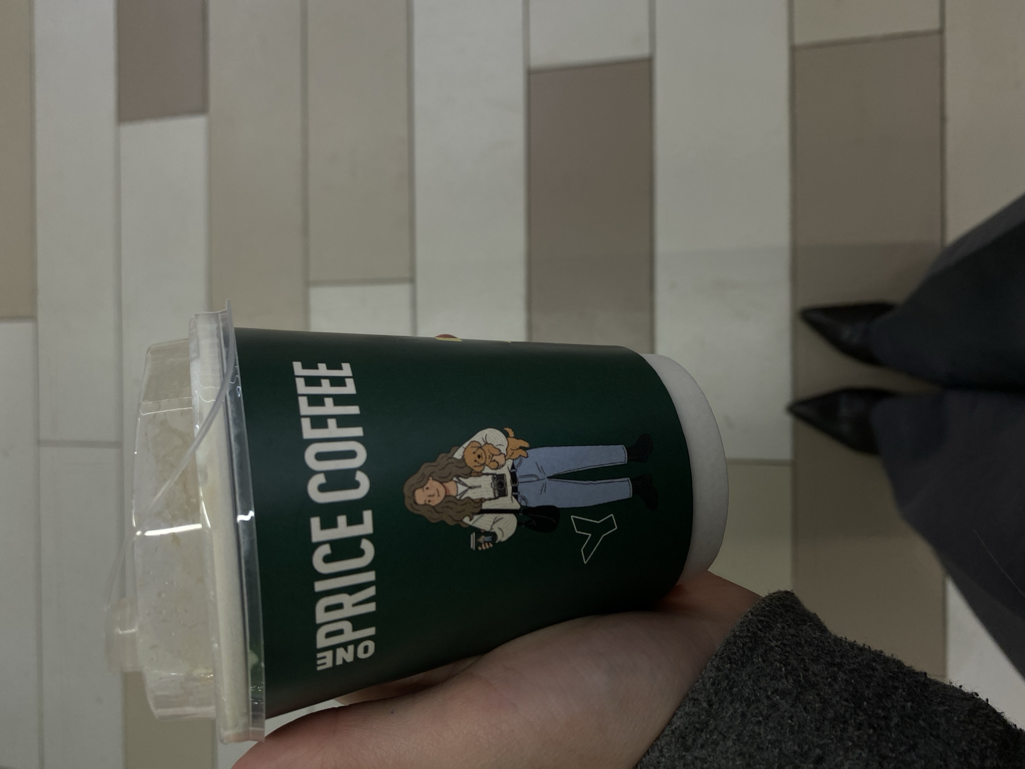 A person holds a cup of "ONE PRICE COFFEE" in their hand. The cup is green with white lettering, and has an illustration of a woman. The drink inside the cup appears to be creamy or light-colored. The background shows a tiled floor and the lower portion of a person wearing dark pants and black shoes. The location is most likely Kazan, Russia, based on the "ONE PRICE COFFEE" branding.