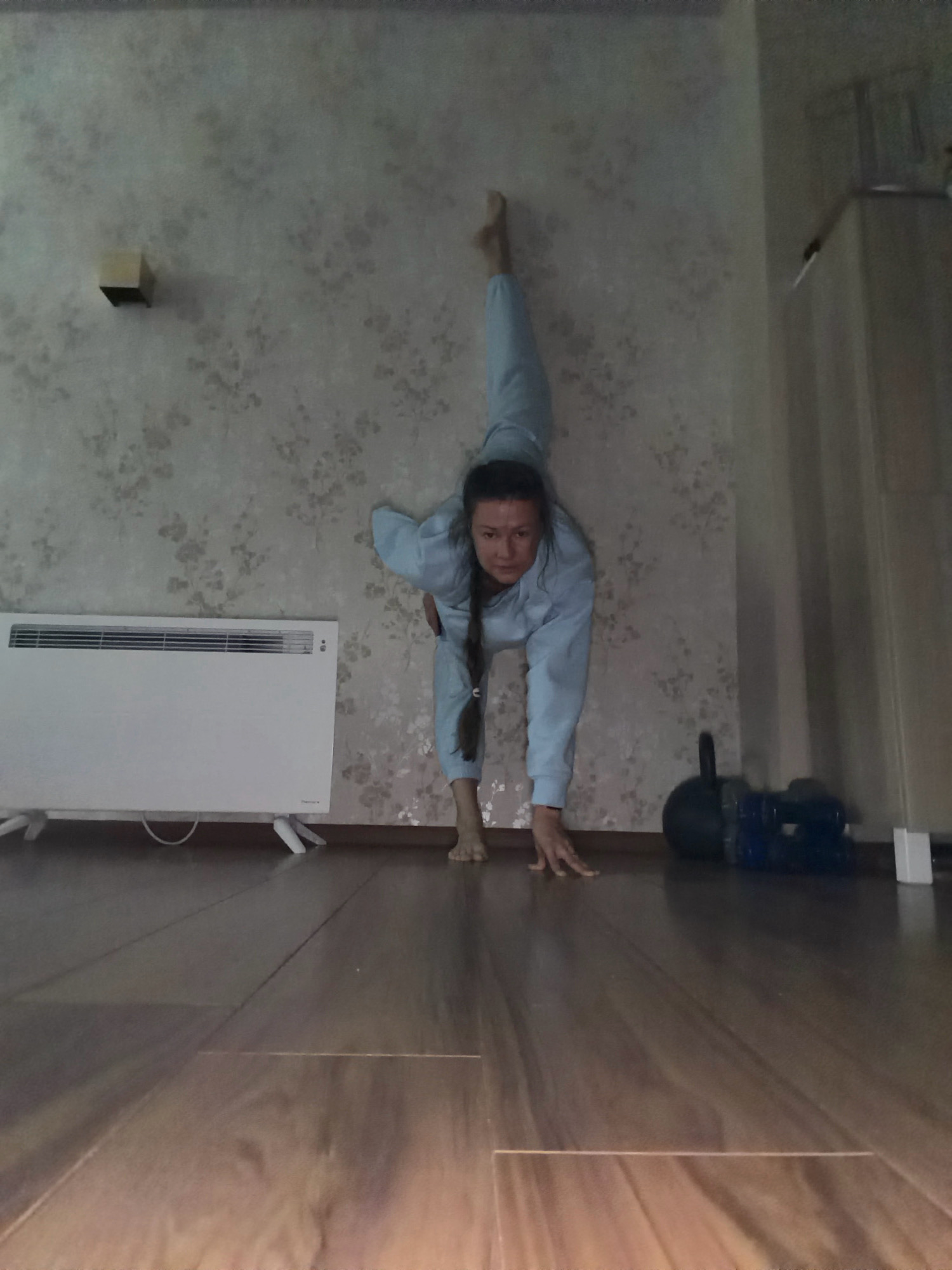 A woman is performing a split against a wall in what appears to be an indoor room. She is wearing a light blue tracksuit and her legs are extended, one upwards and the other bent. Her hands are on the floor for support. To her left, there is a white electric heater. In the background, there are weights and other exercise equipment. The wall has a floral pattern. The floor is made of dark wood planks. The overall setting suggests a home environment. The image was captured in Riga, Latvia.