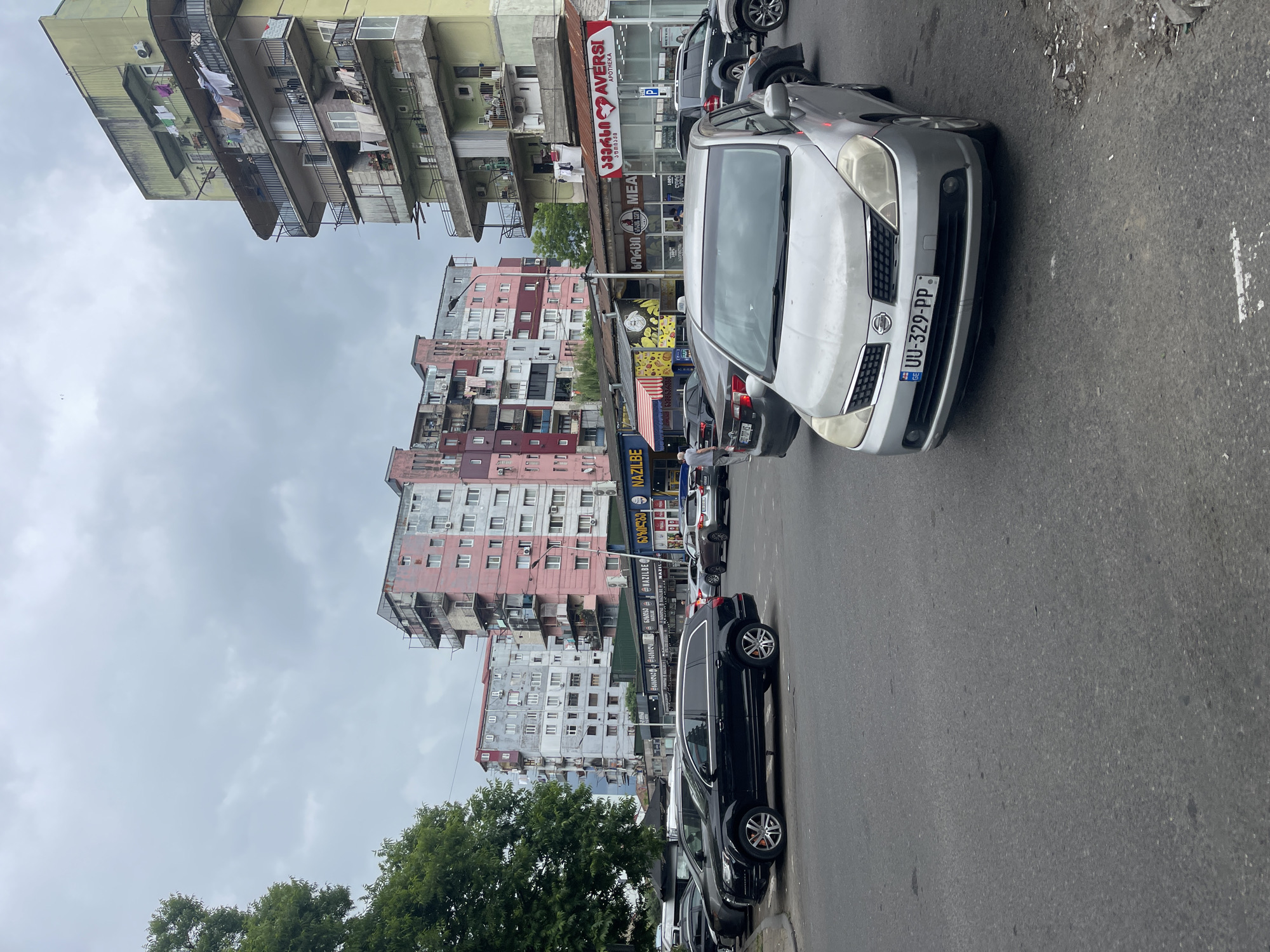 The image depicts a street scene in Batumi, Georgia, characterized by multi-story apartment buildings with visible balconies, some adorned with laundry. In the foreground, a silver Nissan car with license plate UU-329-PP is parked on a paved road. Several other vehicles, including a black SUV and various sedans, are also present in the scene, indicating a populated urban environment. Shops with signage in Georgian script are visible along the street, suggesting commercial activity. The sky is overcast, contributing to a somewhat muted lighting condition. The overall impression is one of a typical urban streetscape in a city with older residential architecture and active commercial areas.