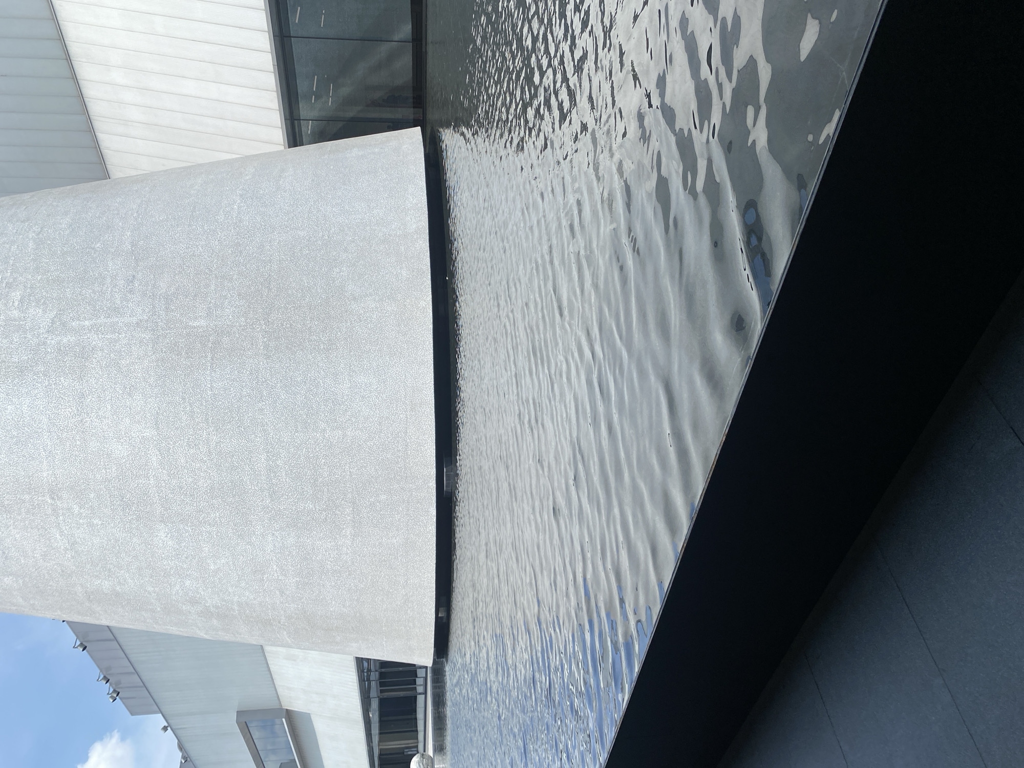 This is an eye-level shot of a modern architectural structure in Bangkok, Thailand. The shot is composed of a reflective pool of water adjacent to a curved, textured gray pillar of the building. The water has light ripples across the surface, reflecting the sky above. The edge of the pool is framed by a dark gray stone. To the left is the rounded pillar, leading to more modern building with glass and metal details. The sky is visible in the lower left corner.