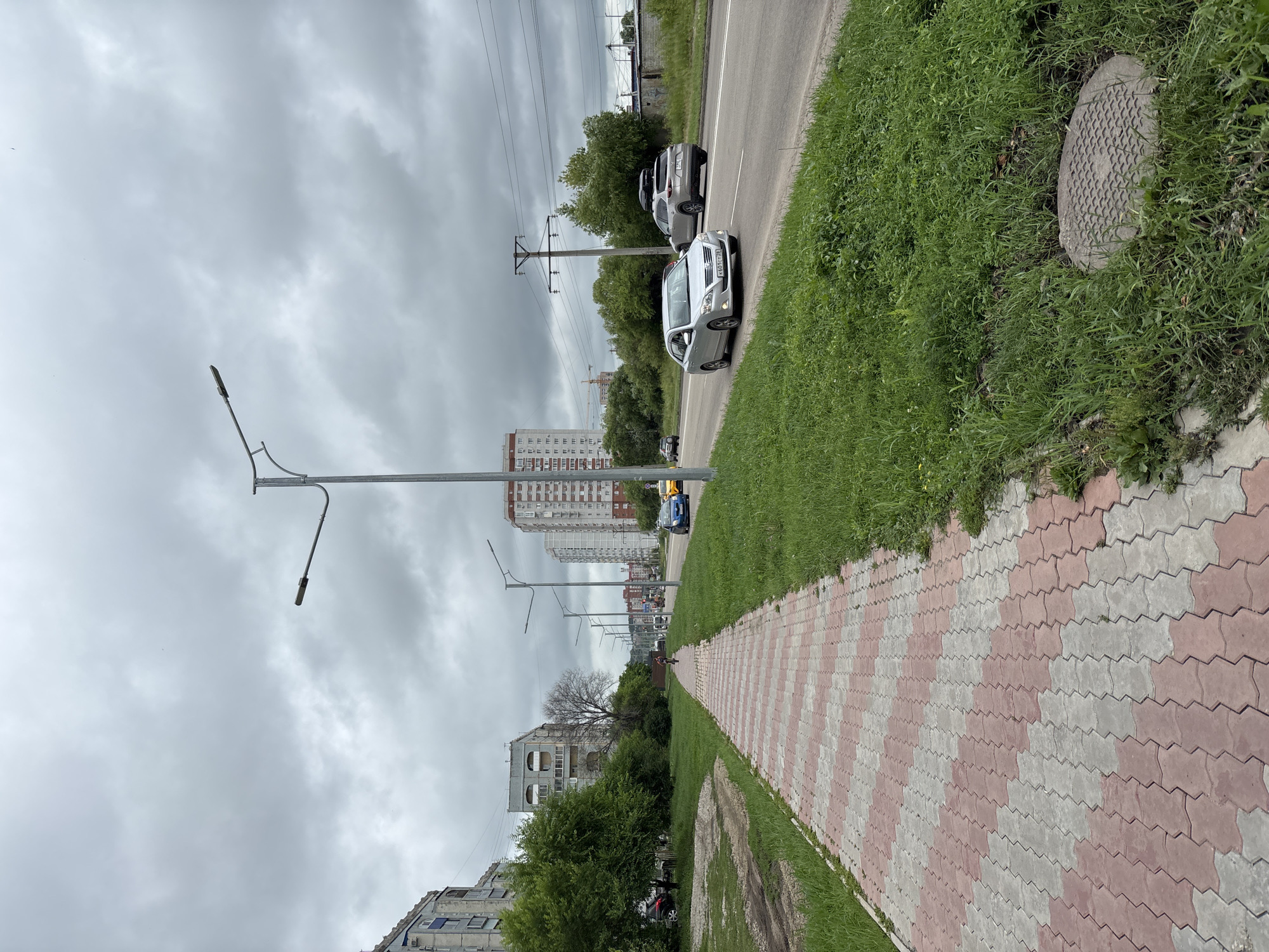 The image depicts a wide, pedestrian pathway paved with red and gray interlocking blocks, leading into the distance along a city street. The pathway is bordered by a strip of green grass. Across the street, there is a multi-lane road with several cars driving, including a silver sedan in the foreground with license plate K651CT 75. Beyond the road, there are buildings, trees, and utility poles under a cloudy sky. To the left of the pathway, there are apartment buildings and green trees. The scene appears to be in Blagoveshchensk, Russia, as indicated by the license plate, which is typical for the Amur Oblast region. A man is riding a bicycle on the pathway in the mid-ground, heading away from the camera.